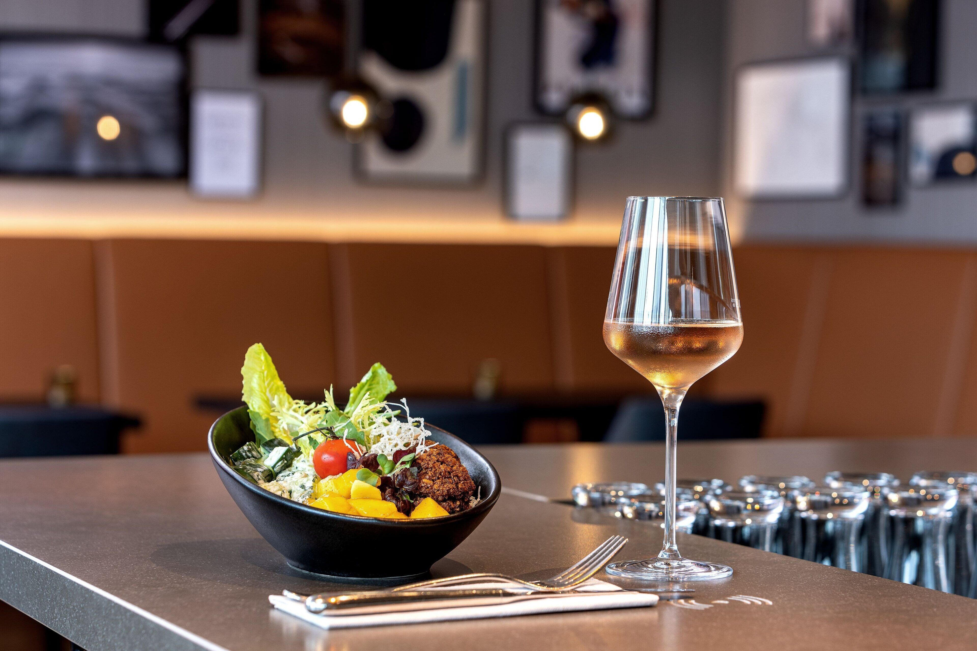A bowl of salad with a glass of white wine on a table in a restaurant.