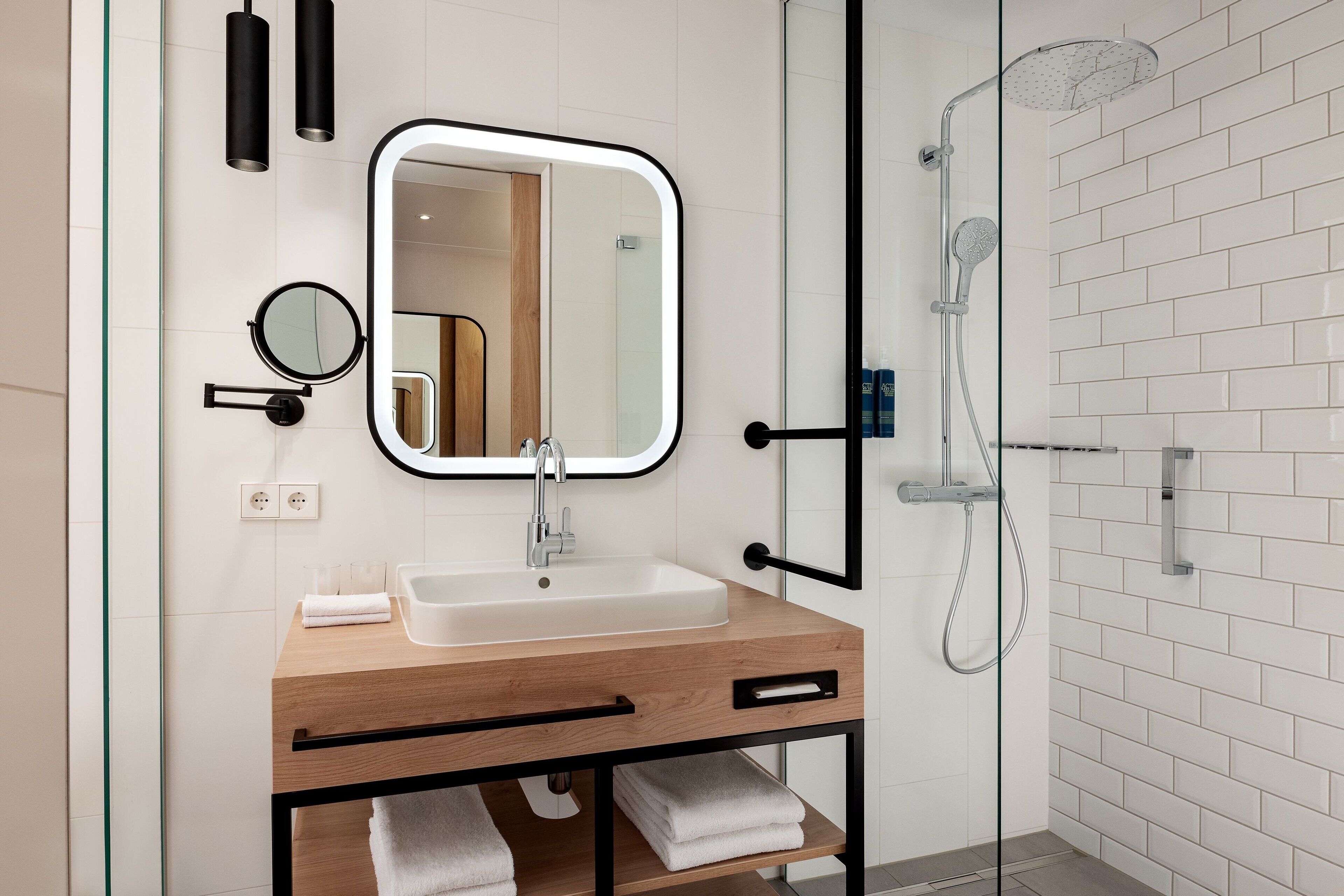 Een moderne badkamer met een houten wastafelmeubel, een ronde spiegel, een douchecabine met glazen wanden en witte betegelde muren.
