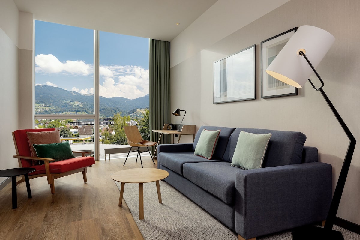 Un salon moderne avec un canapé, un fauteuil et une table basse. Une grande fenêtre offre une vue sur les montagnes et sur le paysage urbain.