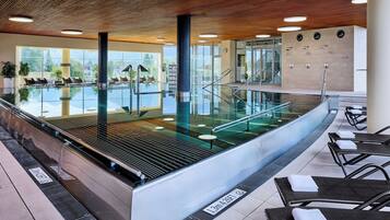 Un área de piscina interior moderna con barandillas de vidrio, sillones de descanso y vista despejada al exterior.