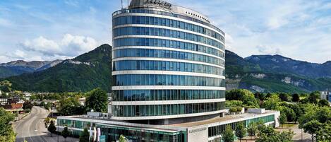 A modern hotel with a curved glass facade, located in a mountainous area with a clear sky.