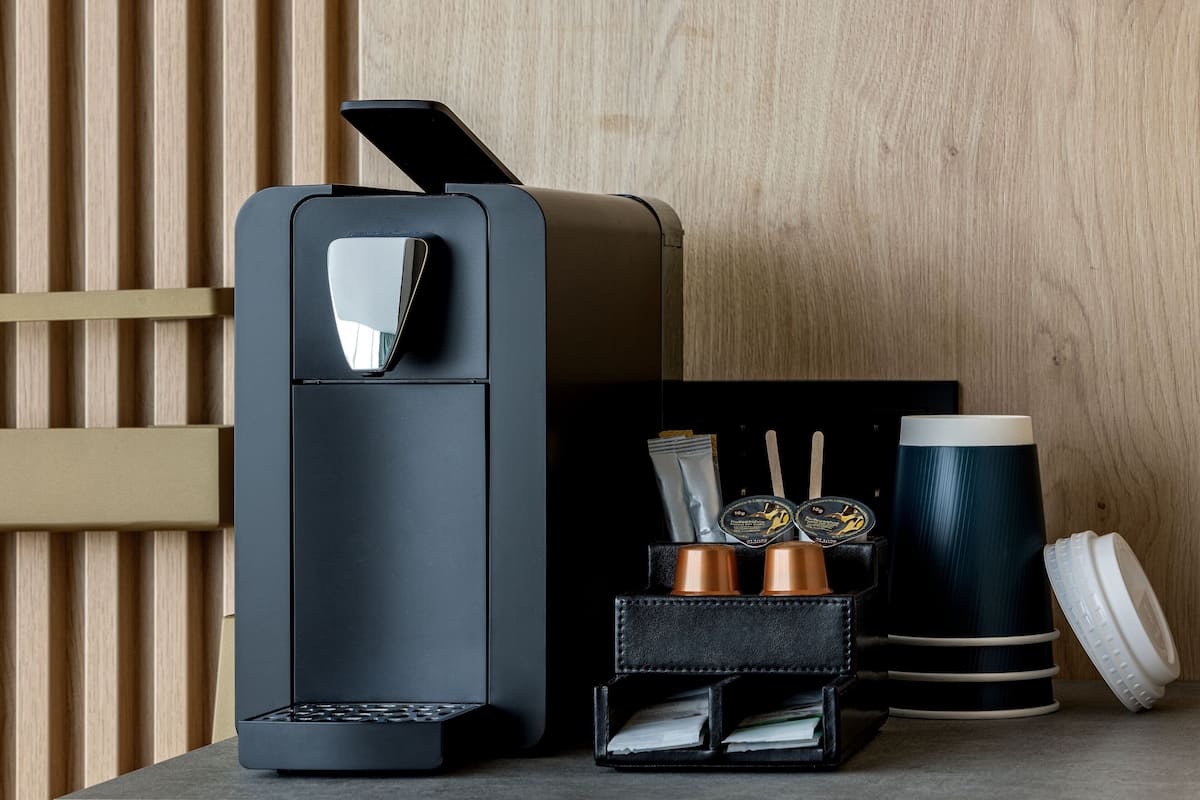 Une machine à café moderne au design épuré, avec une palette de couleurs noir et gris, et un petit plateau contenant une tasse à café, une cuillère et un distributeur de serviettes.