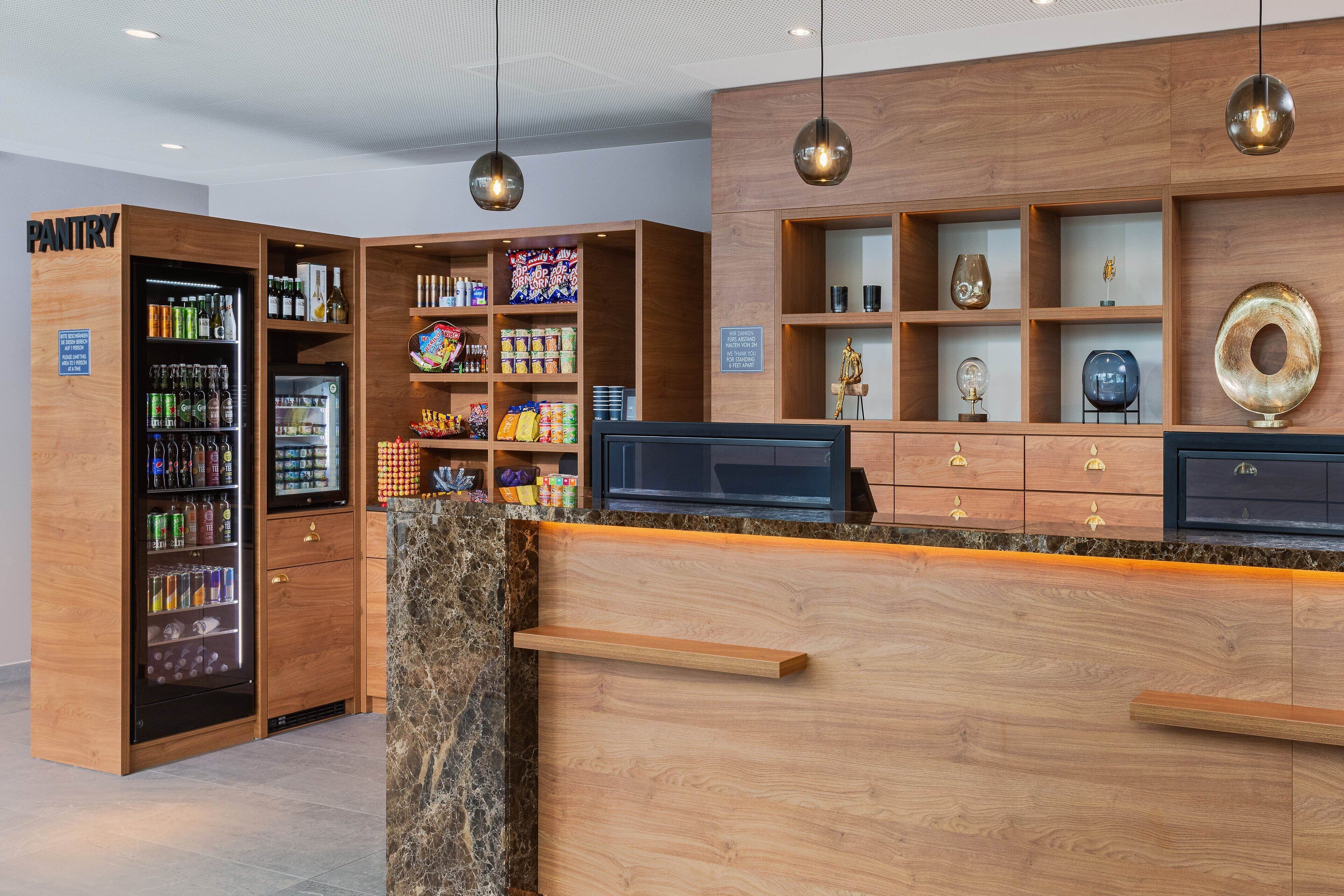 A hotel reception area with a wooden counter, a refrigerated beverage dispenser, and shelves with various items.
