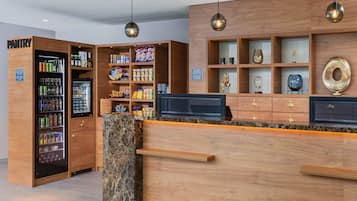 A hotel reception area with a wooden counter, a refrigerated beverage dispenser, and shelves with various items.