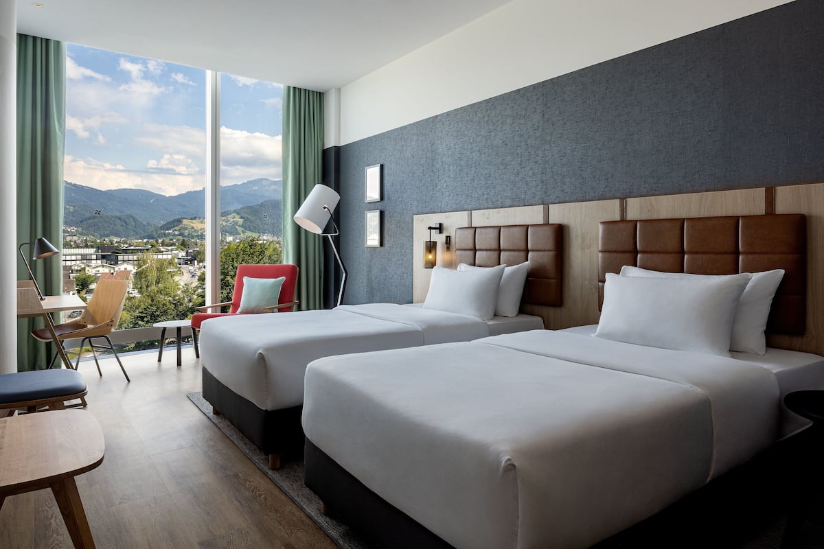 Une chambre d’hôtel avec deux lits, un bureau et une chaise. Une grande fenêtre offre une vue sur les montagnes et sur le paysage urbain.