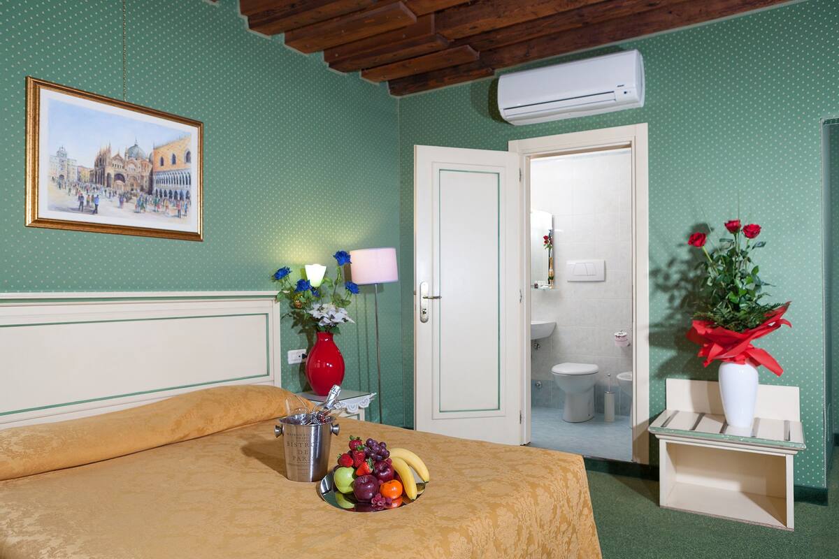 A room with a bed, a dining table with fruit, a vase with flowers, a painting, and a bathroom door.
