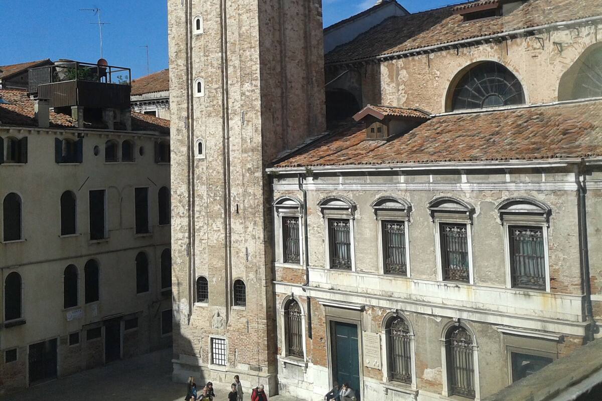 A historic tower with arched windows and a bell, adjacent to a building with a tiled roof and a large arched window.