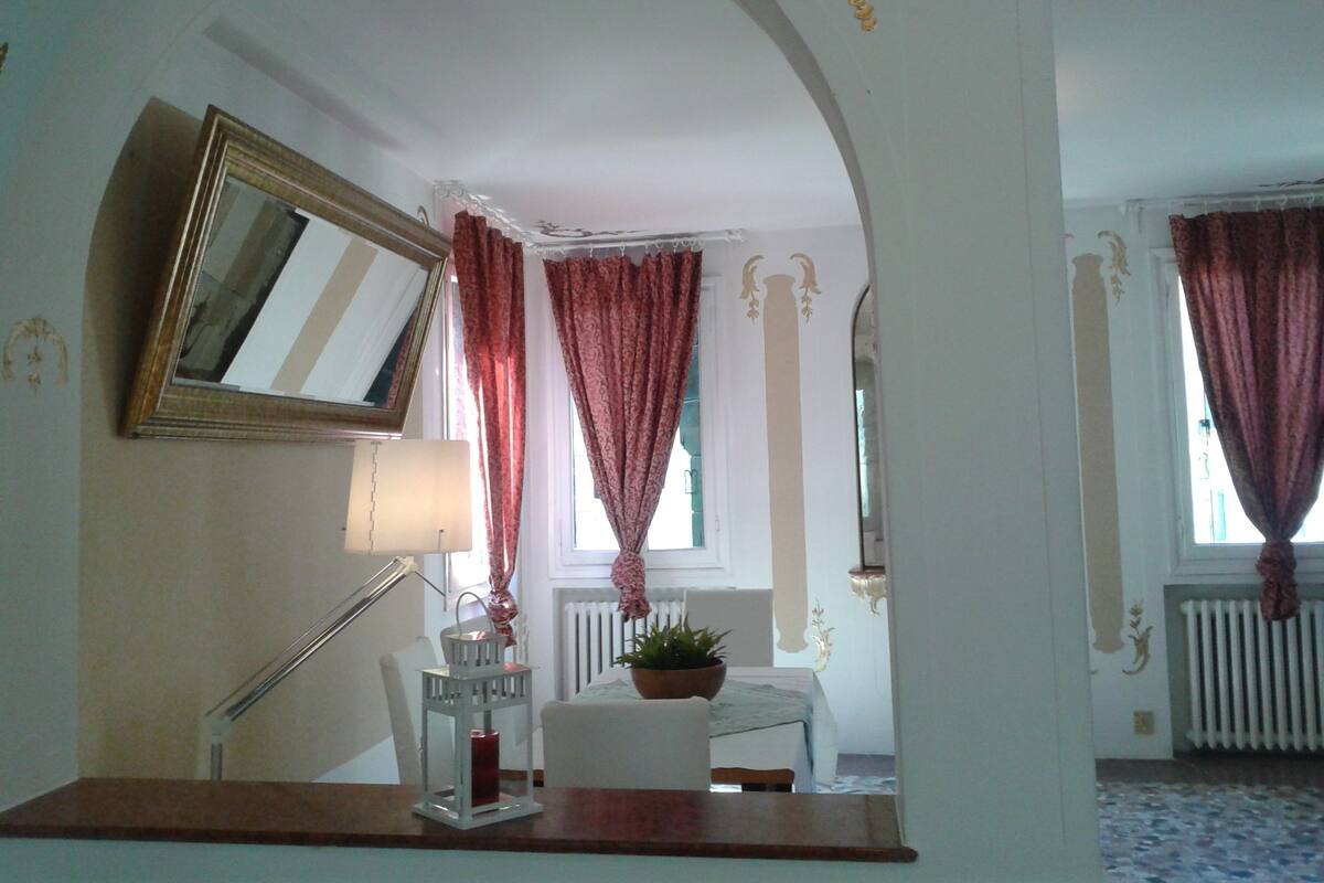 A room with a white arched doorway, a mirror, a lamp, a chair, a table with a candle, and a potted plant.