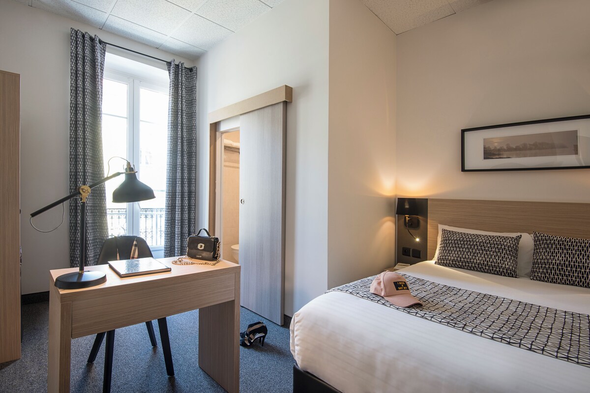 A hotel room with a bed, desk, chair, lamp, and a framed picture on the wall.