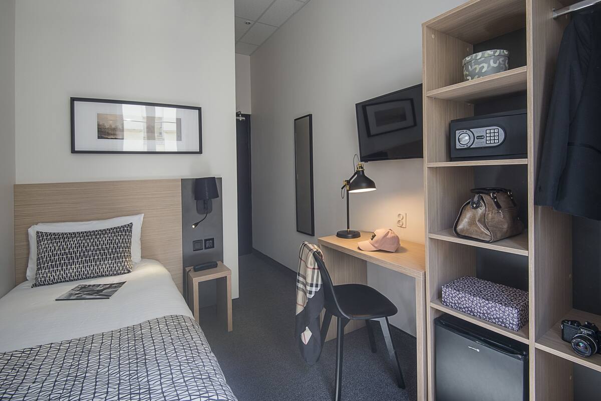 Habitación de hotel con cama, escritorio, silla, estantes y un refrigerador pequeño.