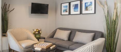 A modern living room with a grey sofa, patterned armchairs, a coffee table with a chess set, a TV mounted on the wall, and framed pictures above the sofa.