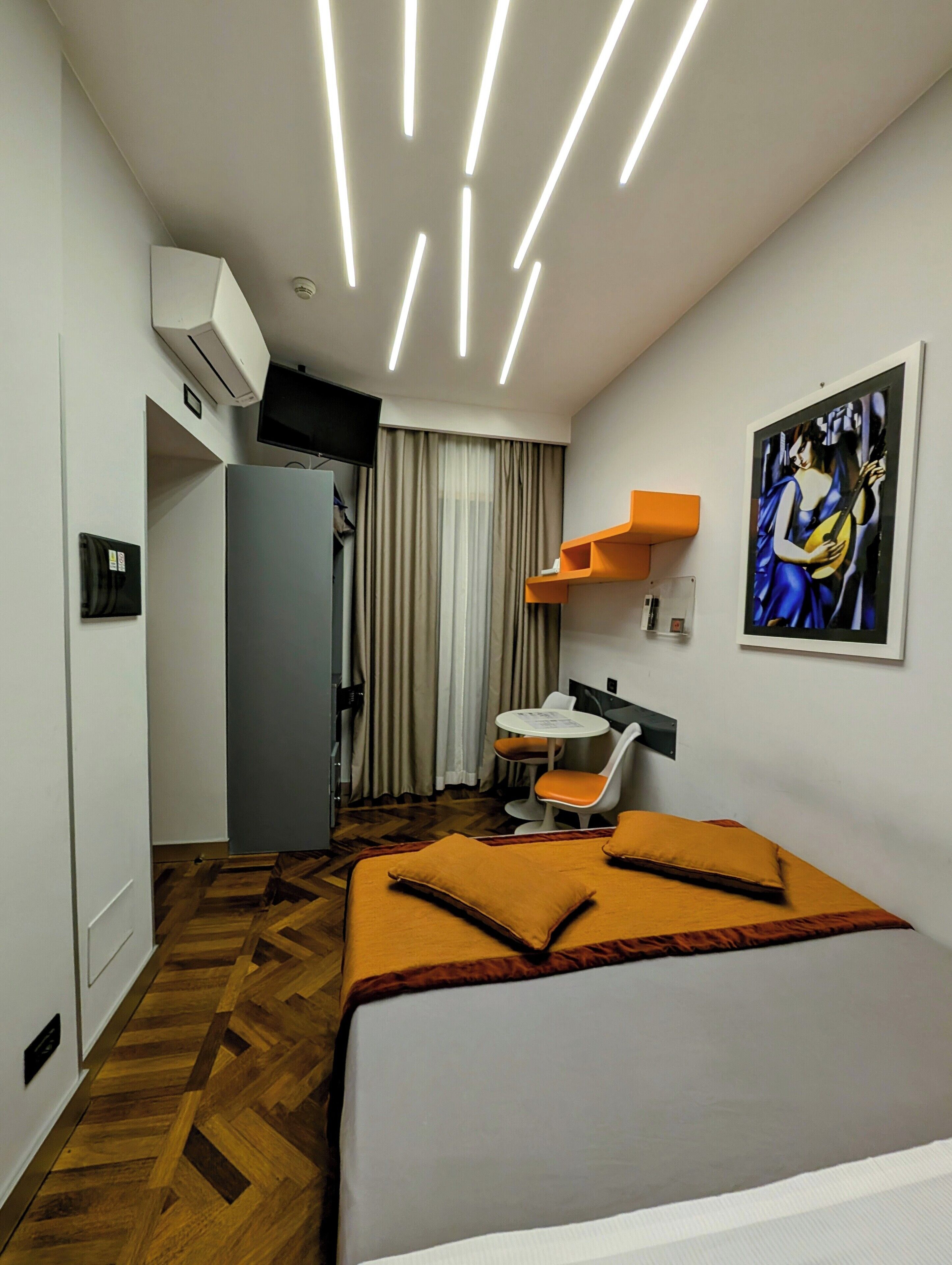 A modern hotel room with a bed, a desk, a chair, and a painting on the wall.