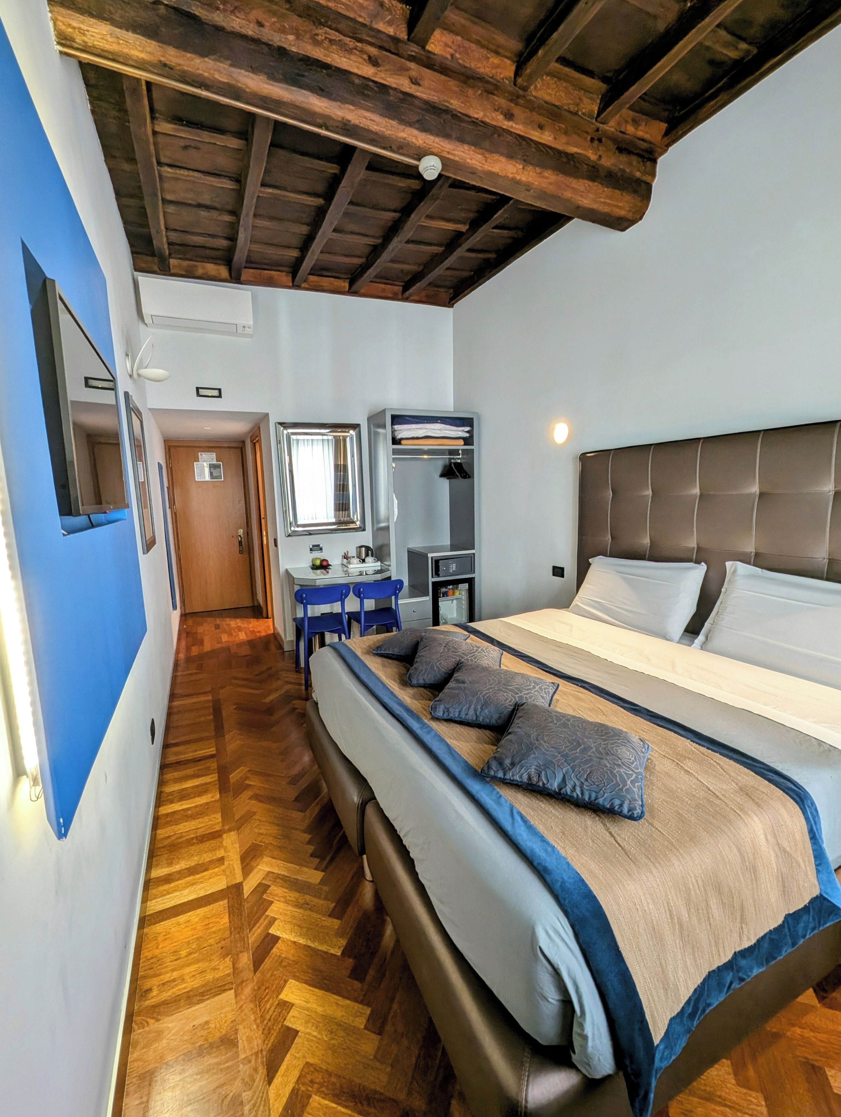 A hotel room with a wooden herringbone floor, a large bed with a headboard, a desk with a chair, a wardrobe, and a mirror.