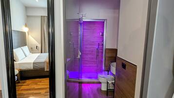 A modern hotel room with a wooden floor, a bed with white linens, a glass shower enclosure, and a bathroom with a toilet and sink.