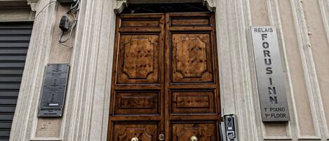 A large wooden double door with a brass doorknob, flanked by two windows with shutters, and a sign indicating the building is on the first floor.
