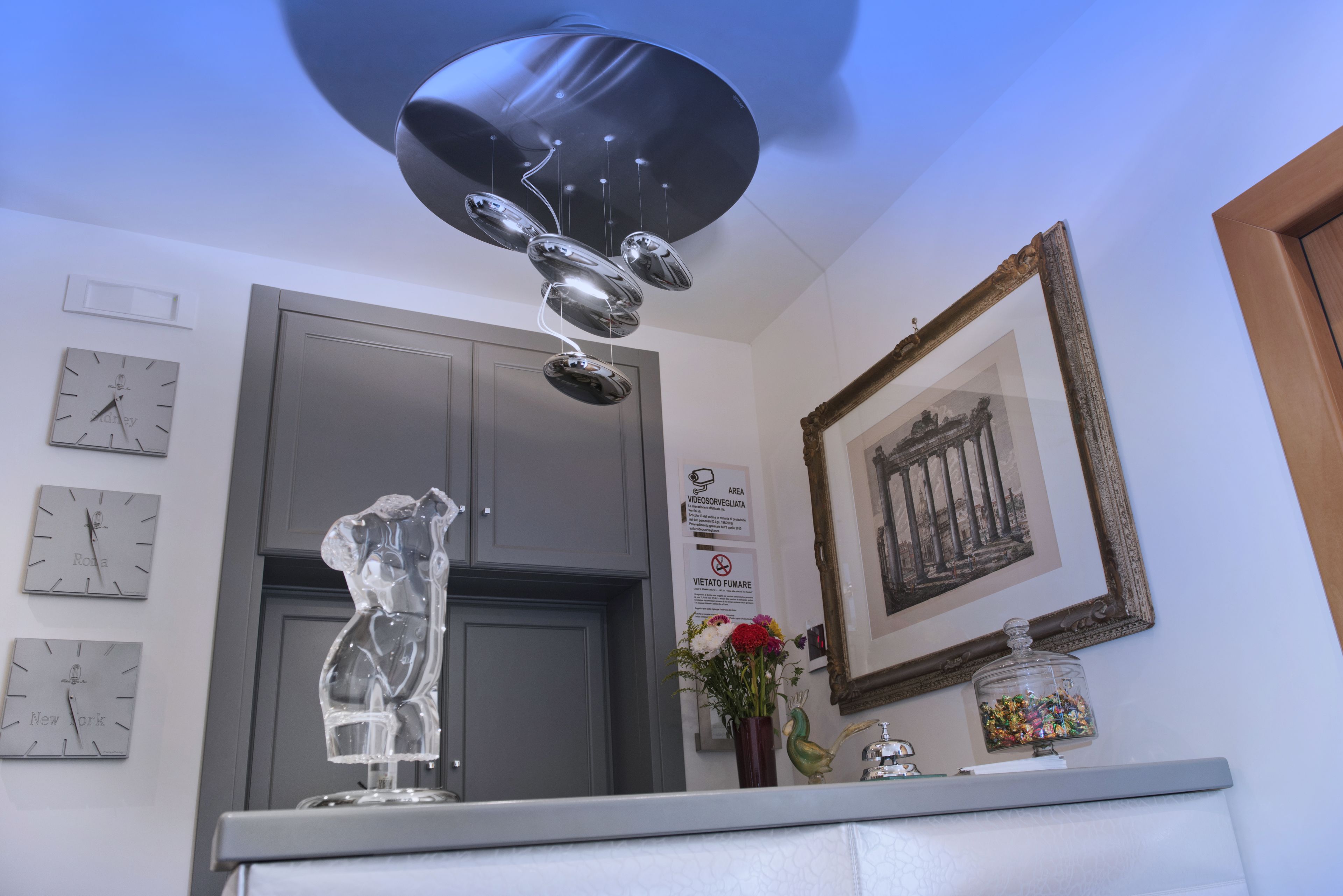 A room with a modern chandelier, a decorative glass sculpture, a framed picture, and clocks on the wall.