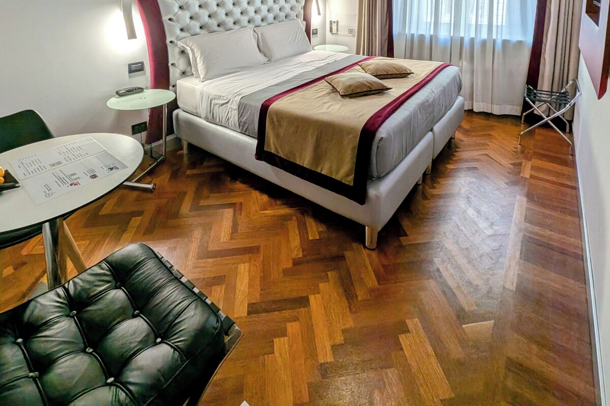 Une chambre avec un plafond en bois, un lit doté d’une tête de lit capitonnée, une table de chevet, une chaise et une fenêtre avec des rideaux.