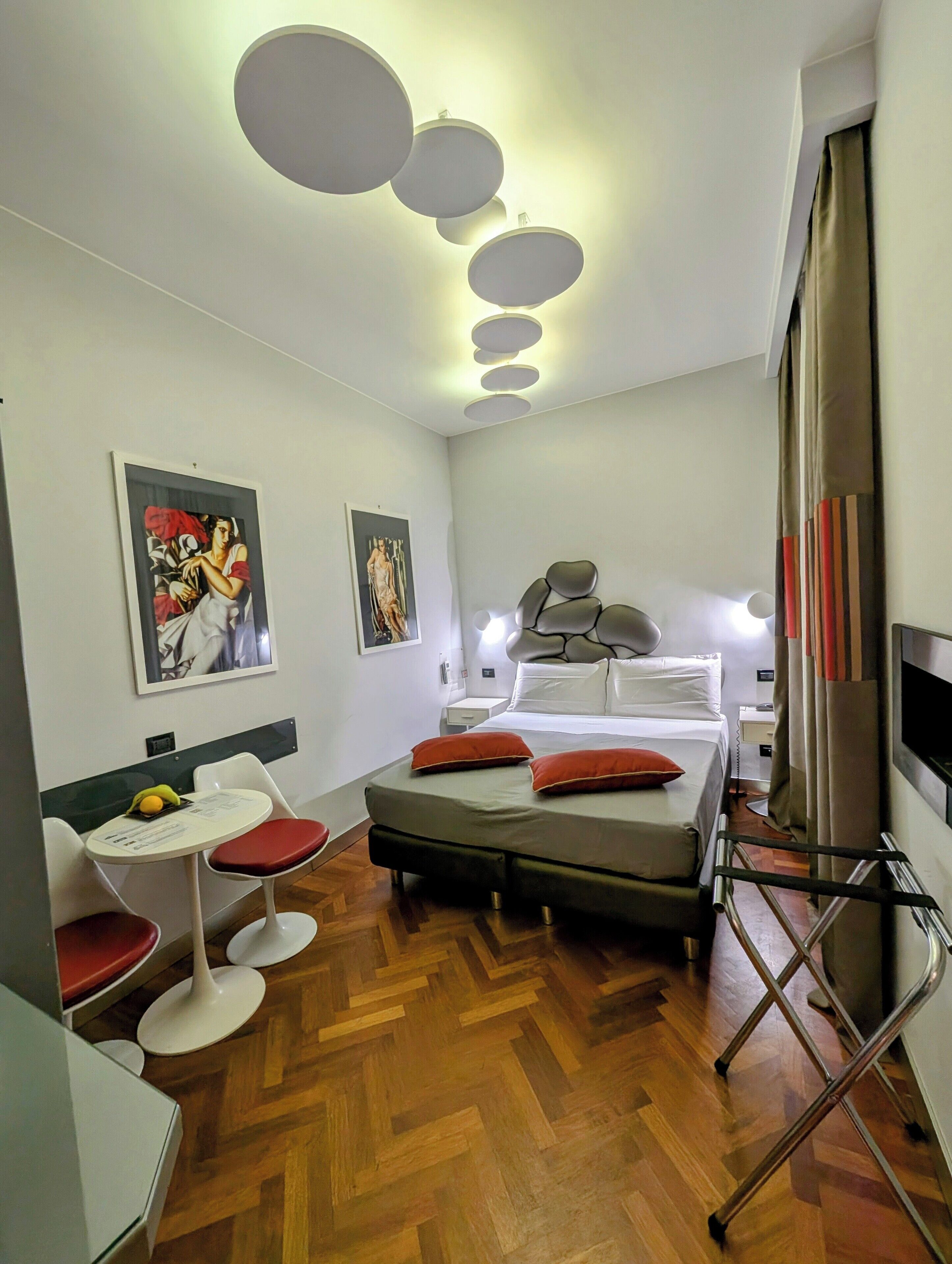 Une chambre d’hôtel moderne, équipée d’un lit, de tables de chevet, d’une petite table ronde avec des chaises, d’œuvres d’art accrochées aux murs et d’un luminaire au plafond.