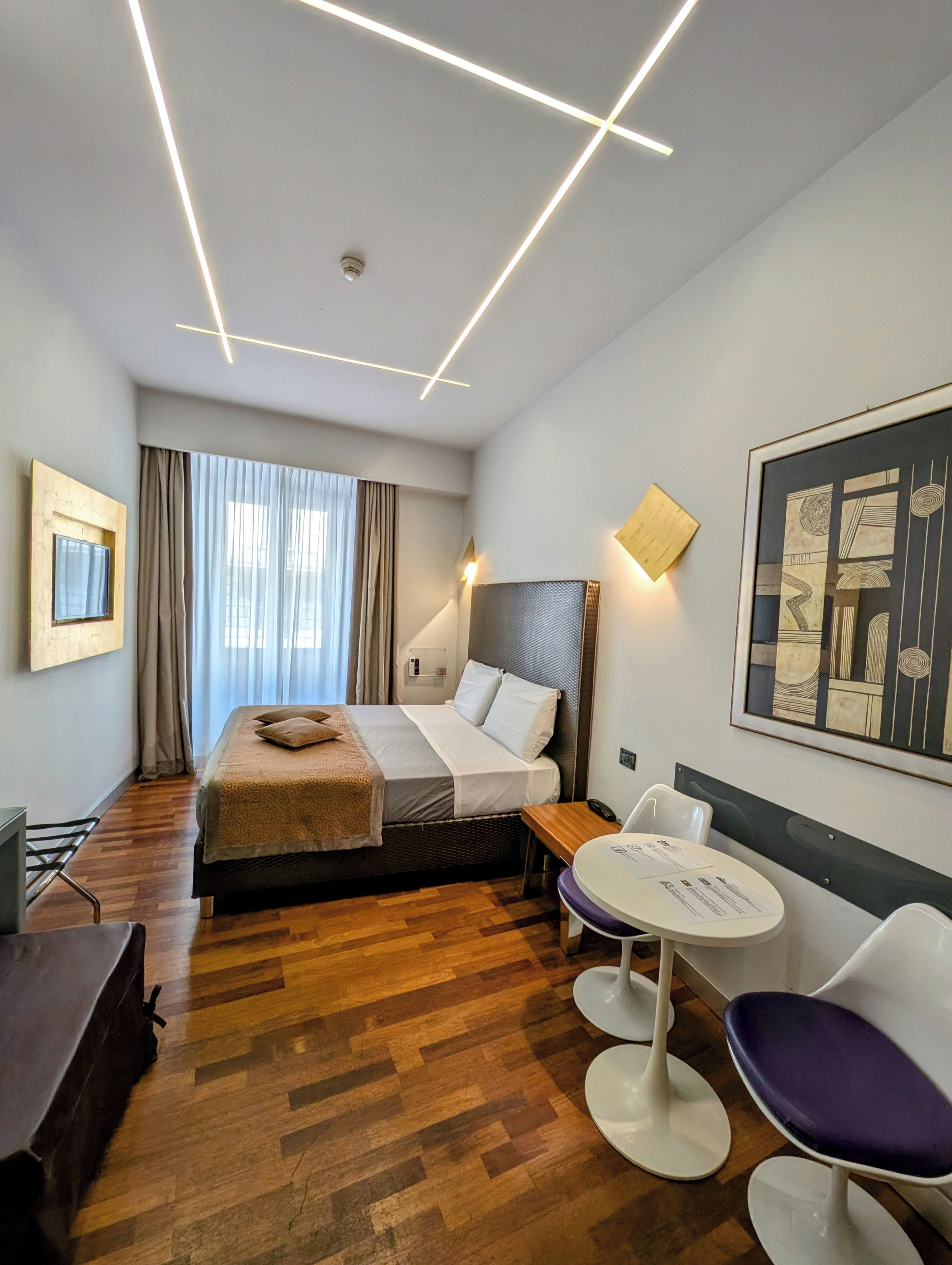 A modern hotel room with a wooden floor, a bed with a headboard, a desk with a chair, a round table with a chair, and a large abstract painting on the wall.