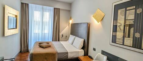 A modern hotel room with a wooden floor, a bed with a headboard, a desk with a chair, a round table with a chair, and a large abstract painting on the wall.