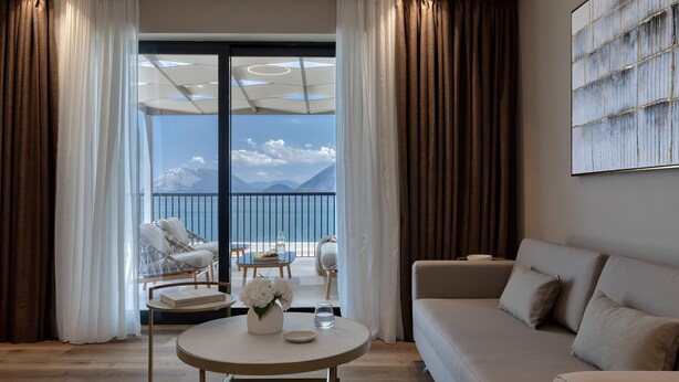 A modern living room with a sofa, a round table with a vase and a glass, and a view of mountains through a large window. - Airotel Achaia Beach (Patras)