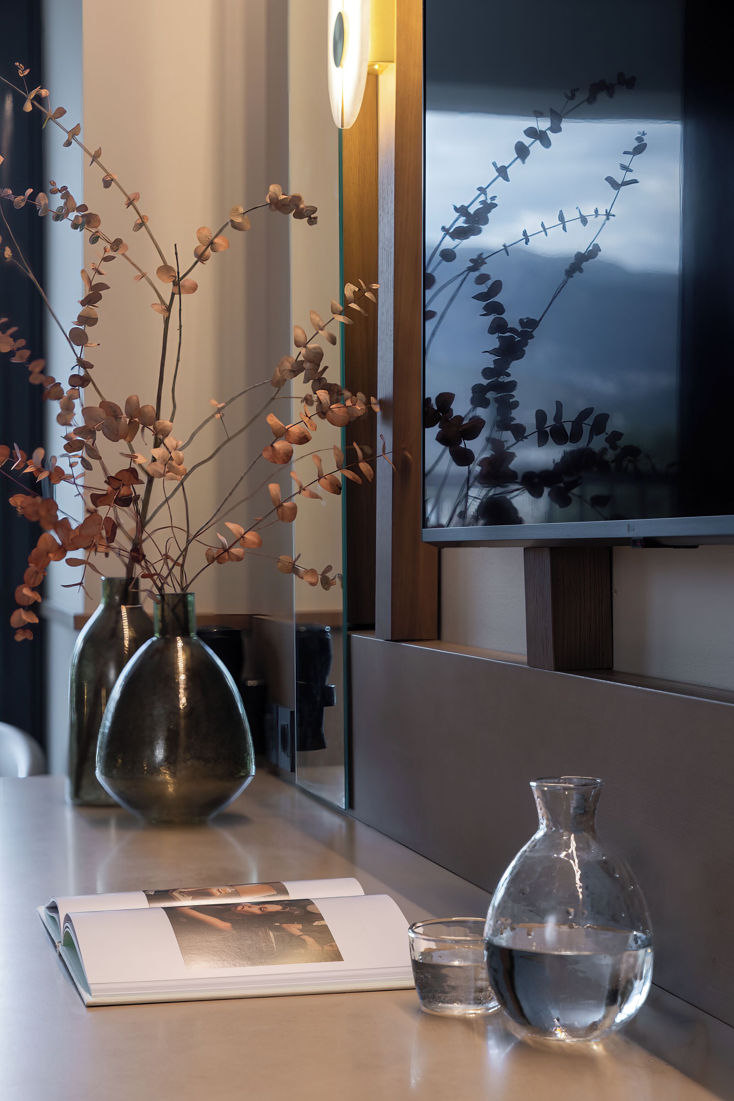 A modern interior with a vase of dried branches, a clear glass vase with water, and a magazine on a table.
