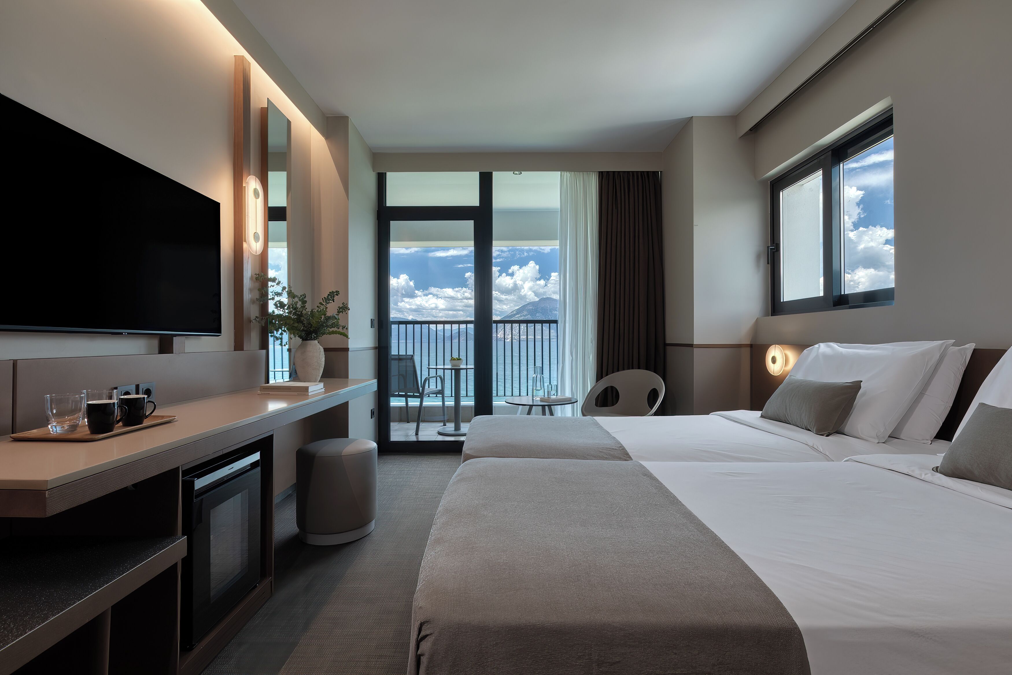 Hotelkamer met flatscreen televisie, bureau met vaas, twee bedden, balkon met uitzicht op de lucht en wolken, en een stoel.