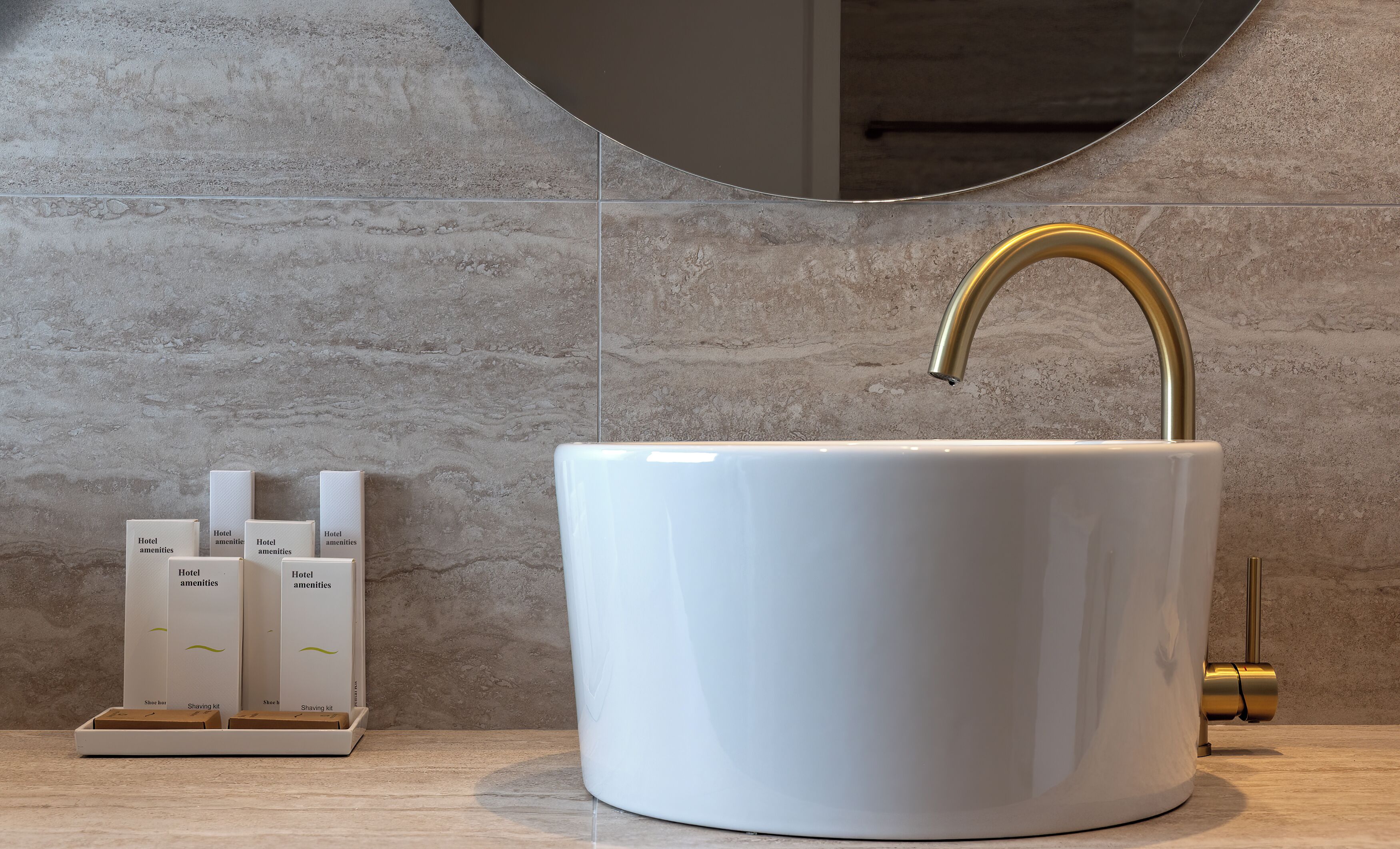 A bathroom with a white sink, gold faucet, and a mirror above it. There are three hotel amenities boxes on the counter.