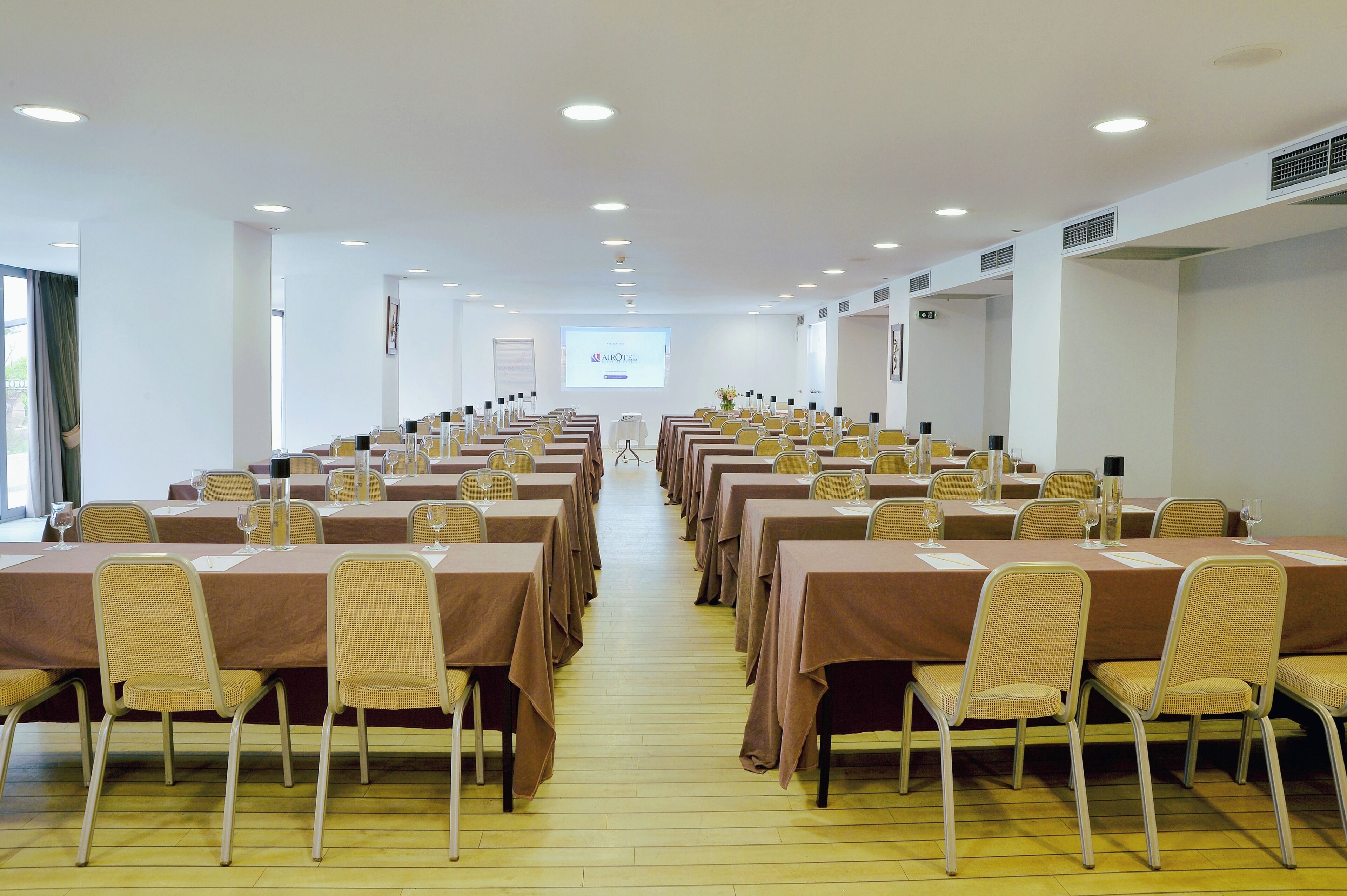 A conference room with rows of tables and chairs, a projector screen, and a window with curtains.