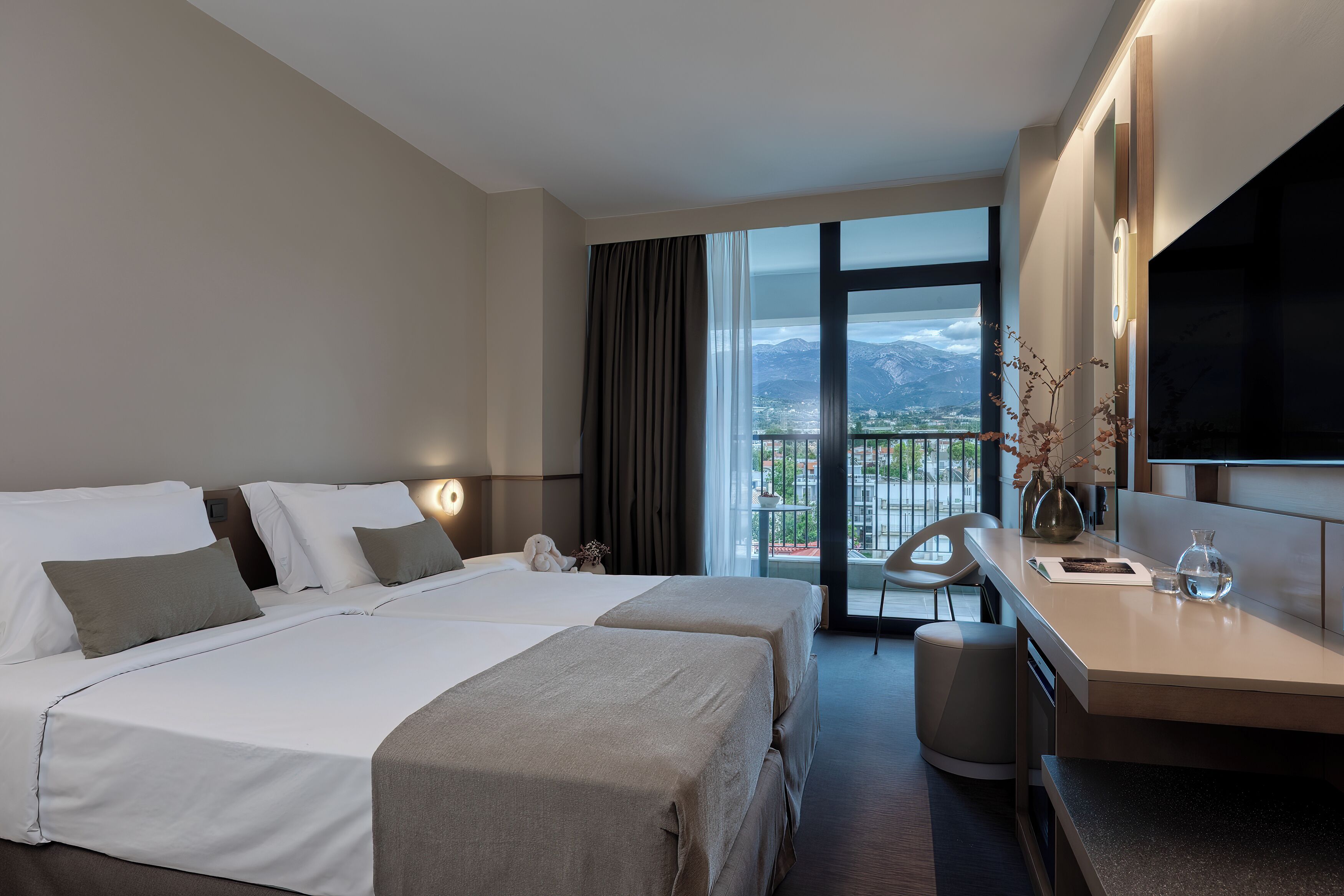 A hotel room with two beds, a desk, a chair, a TV, and a balcony with a mountain view.