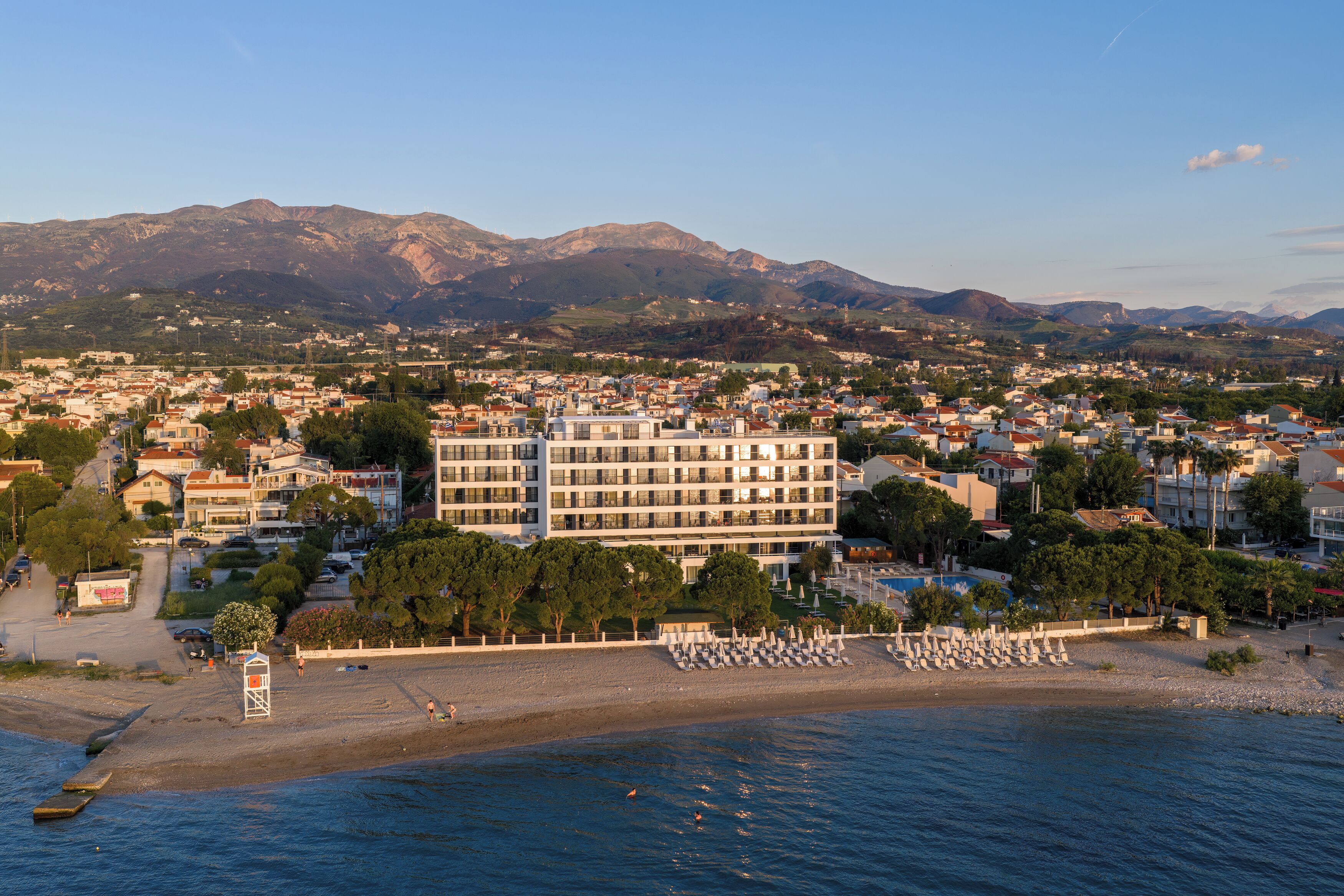 Paisagem costeira com um hotel imponente, vários andares e varandas, uma praia com espreguiçadeiras e montanhas ao fundo.