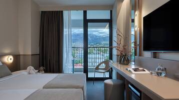 Ein Hotelzimmer mit zwei Betten, einem Schreibtisch, einem Stuhl, einem Fernseher und einem Balkon mit Bergblick.