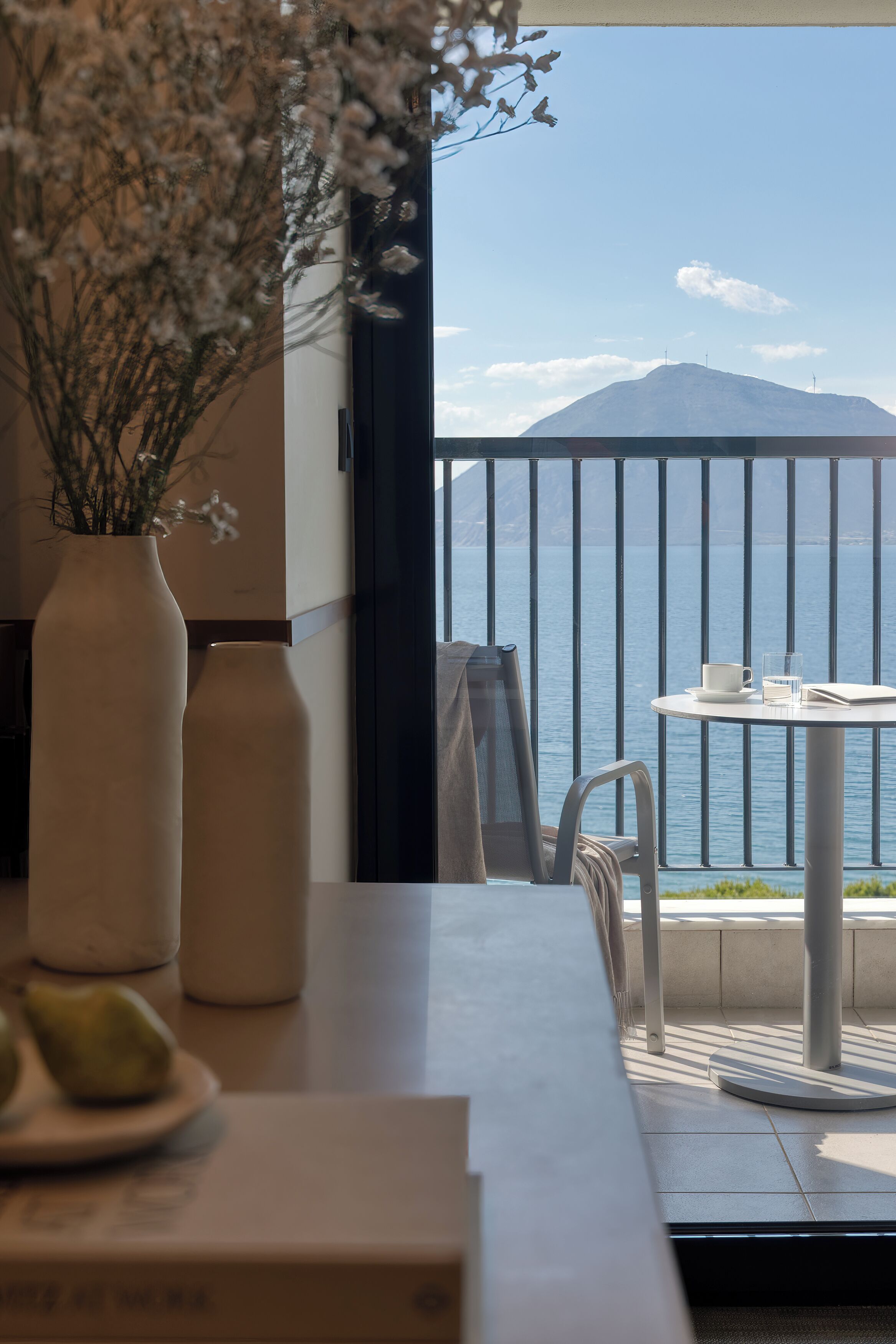 Un balcon avec vue sur la mer et une montagne, une table avec une tasse et un verre, et deux vases remplis de fleurs.