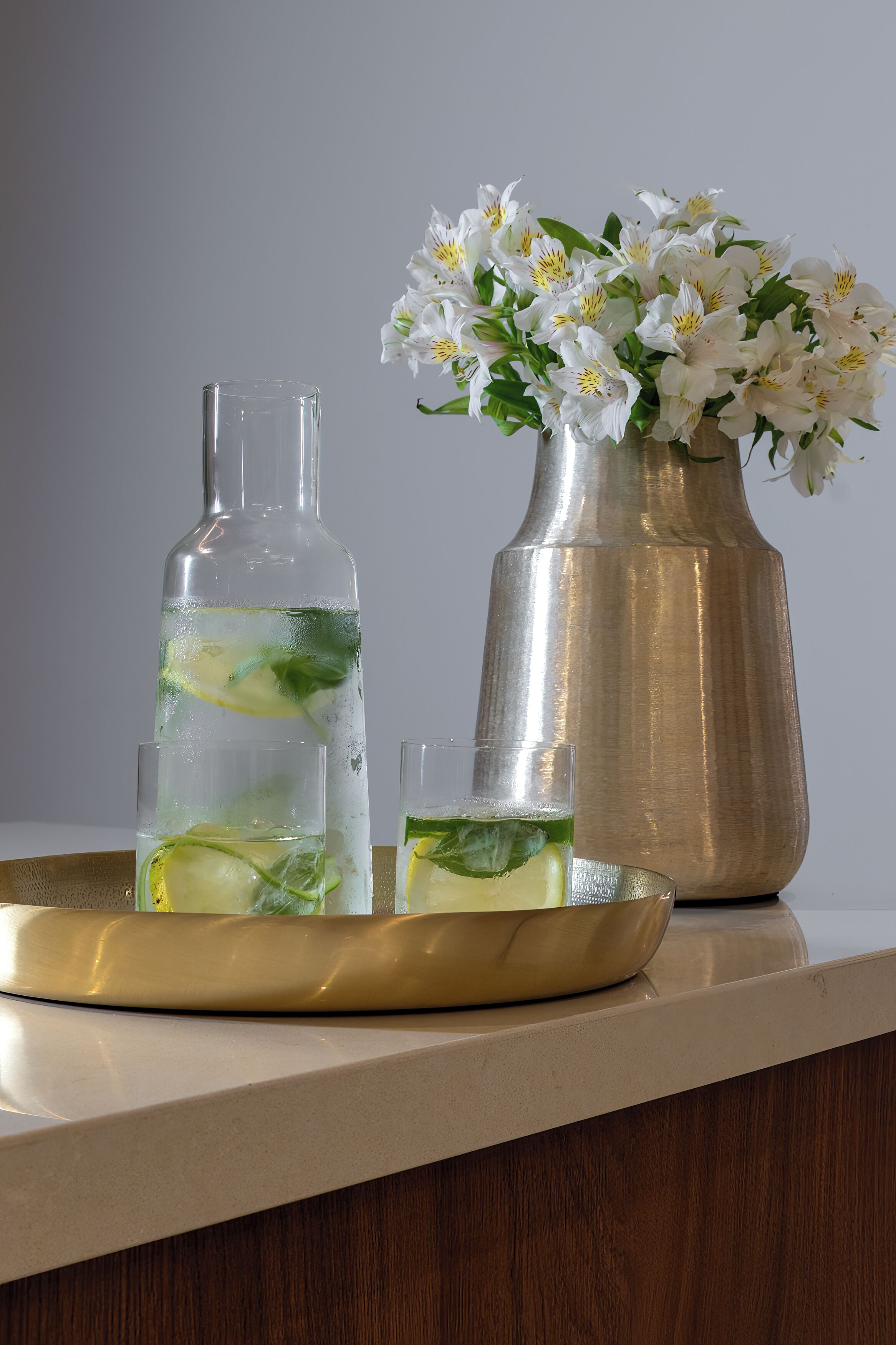 Una botella de vidrio transparente, un florero con flores blancas y dos vasos sobre una bandeja.