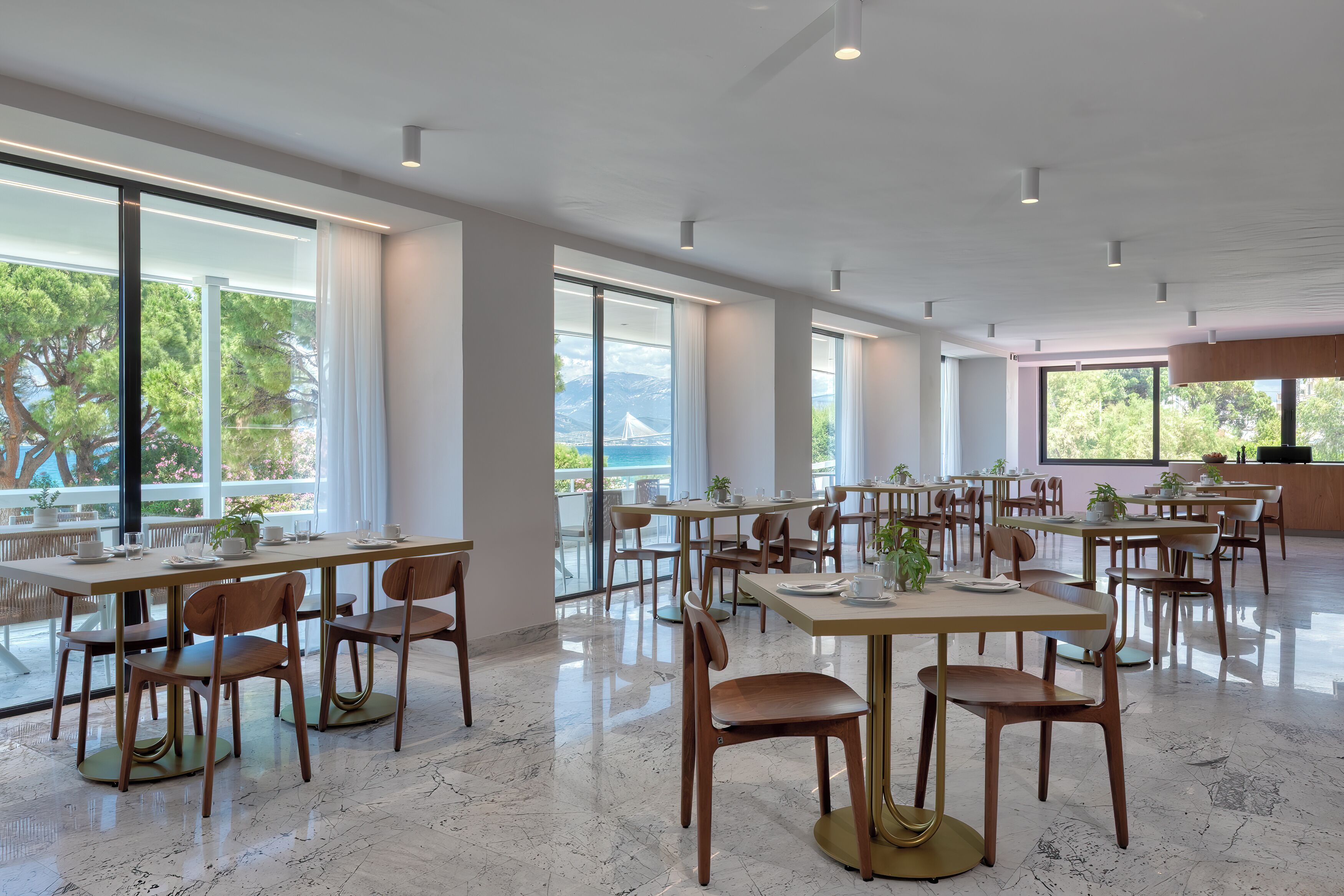Un comedor amplio con varias mesas y sillas, ventanas grandes con vistas a la vegetación y un puente, y un diseño interior moderno y cuidado.