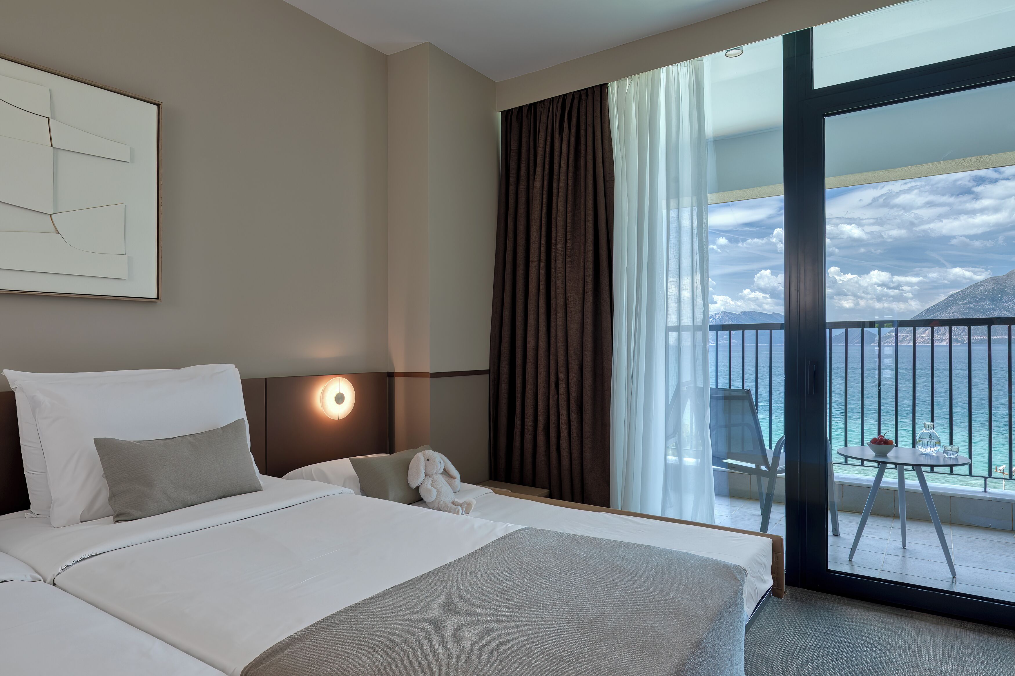 A hotel room with a bed, a nightstand, a lamp, a window with curtains, a balcony with a table and chairs, and a view of the sea and mountains.