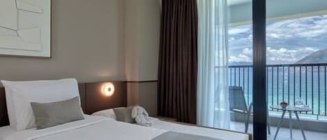 A hotel room with a bed, a nightstand, a lamp, a window with curtains, a balcony with a table and chairs, and a view of the sea and mountains.