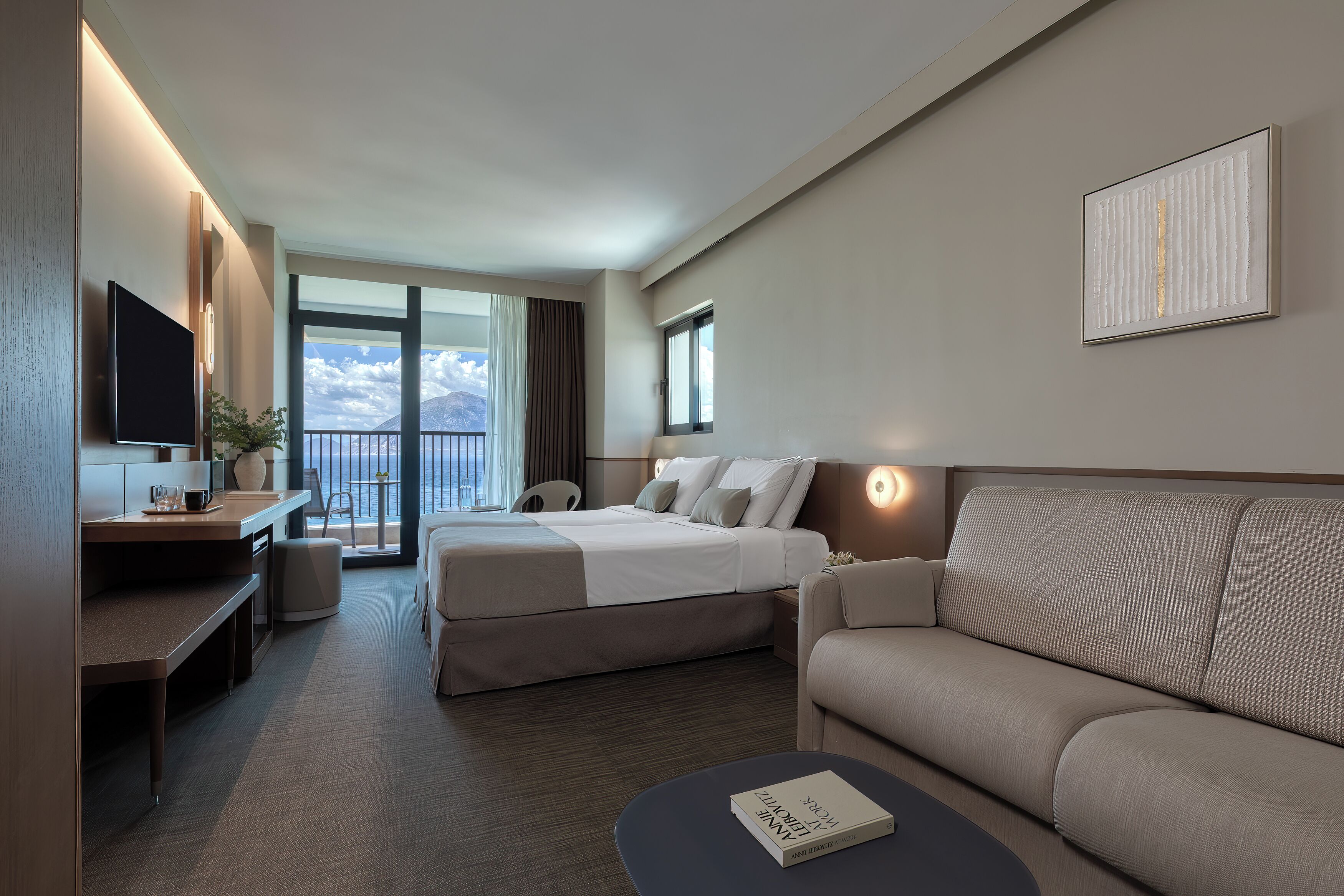 A hotel room with a bed, a sofa, a desk with a flat-screen TV, a bedside table, and a large window with a view of the sky.
