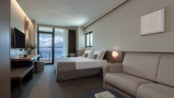 A hotel room with a bed, a sofa, a desk with a flat-screen TV, a bedside table, and a large window with a view of the sky.