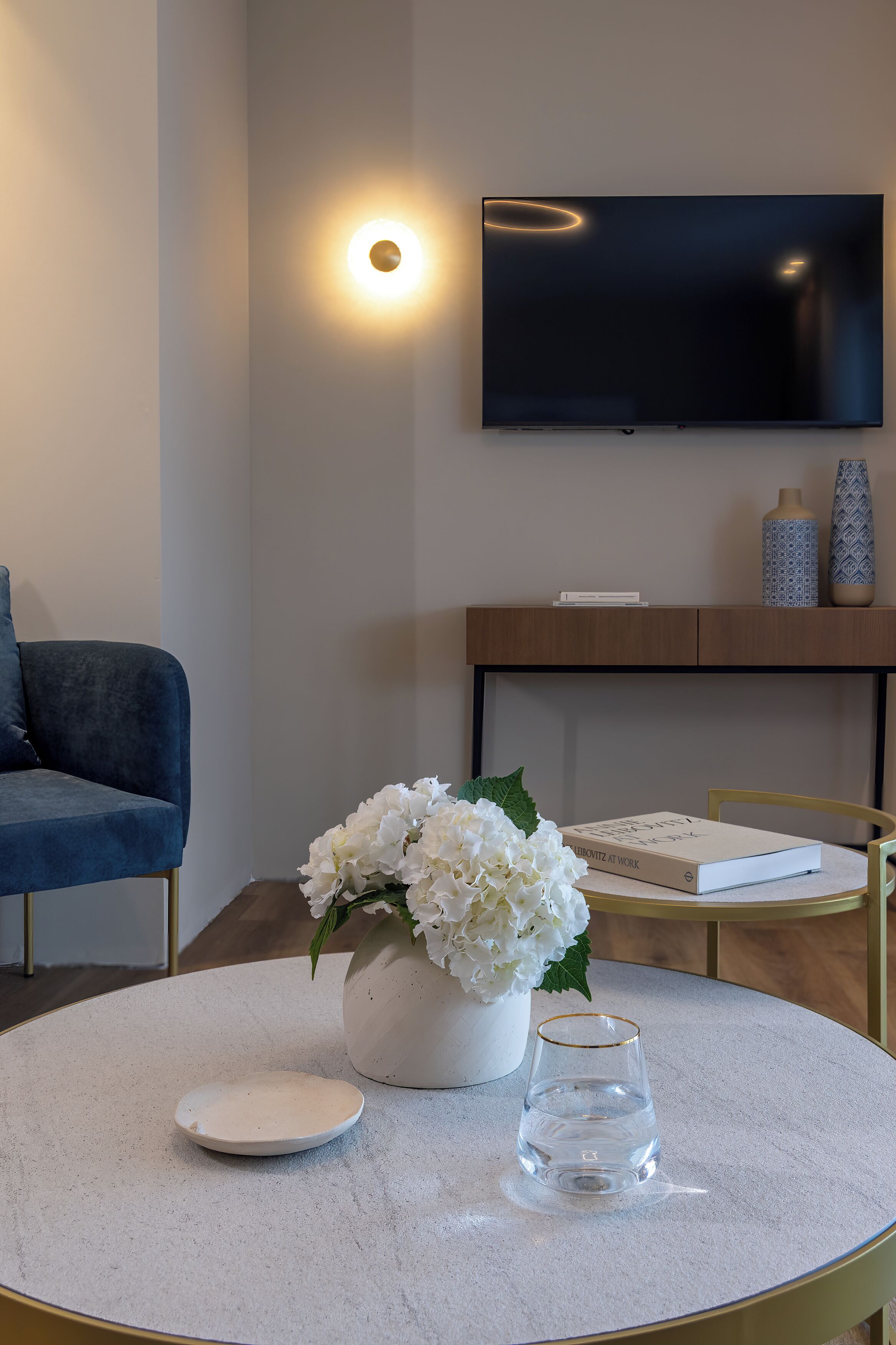 A modern living room with a flat-screen TV, a round table with a vase of flowers, a book, and a glass of water.