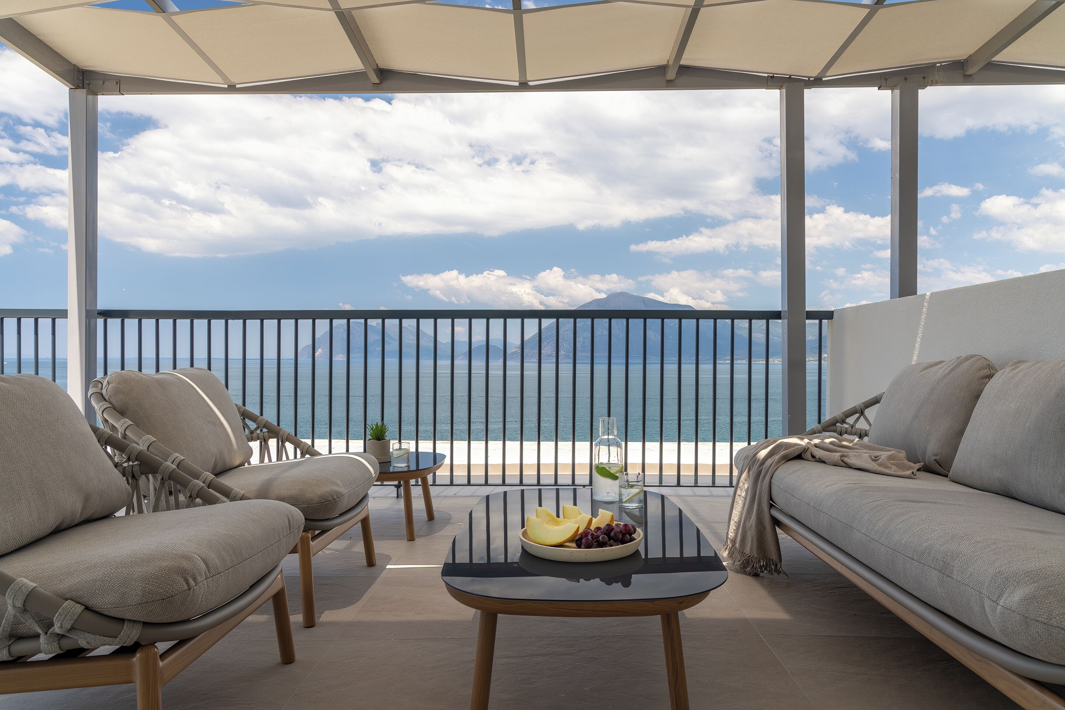 A balcony with a view of the sea, a sofa, a table with fruit, and a small table with a plant.