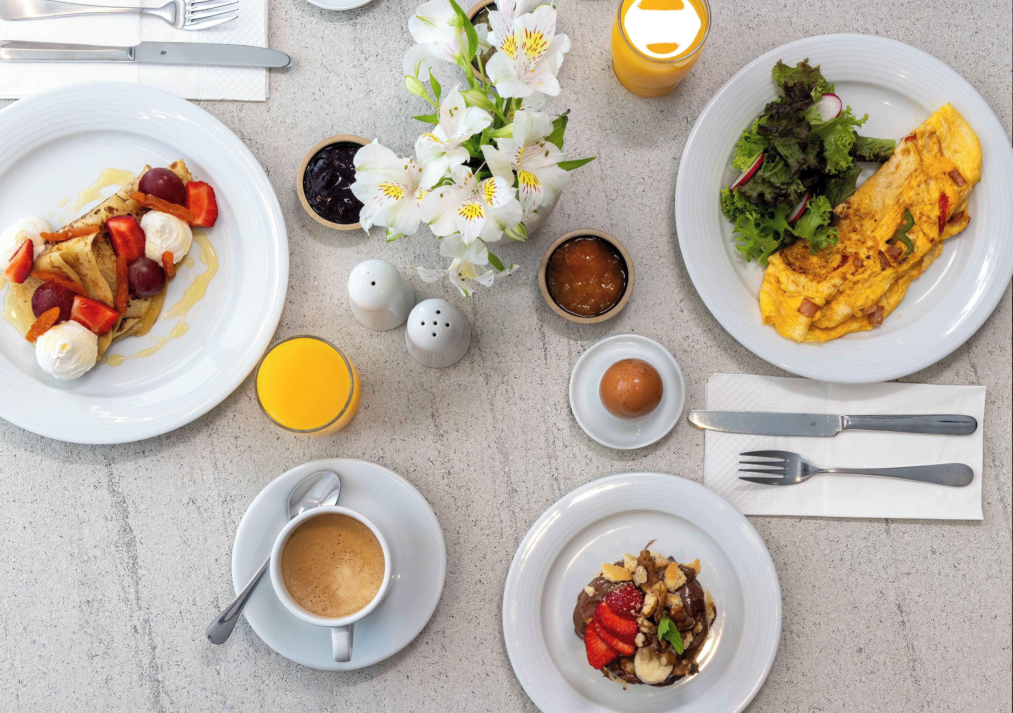 En frukostuppdukning med tallrikar fyllda med mat, en kopp kaffe, ett glas apelsinjuice och en vas med blommor.