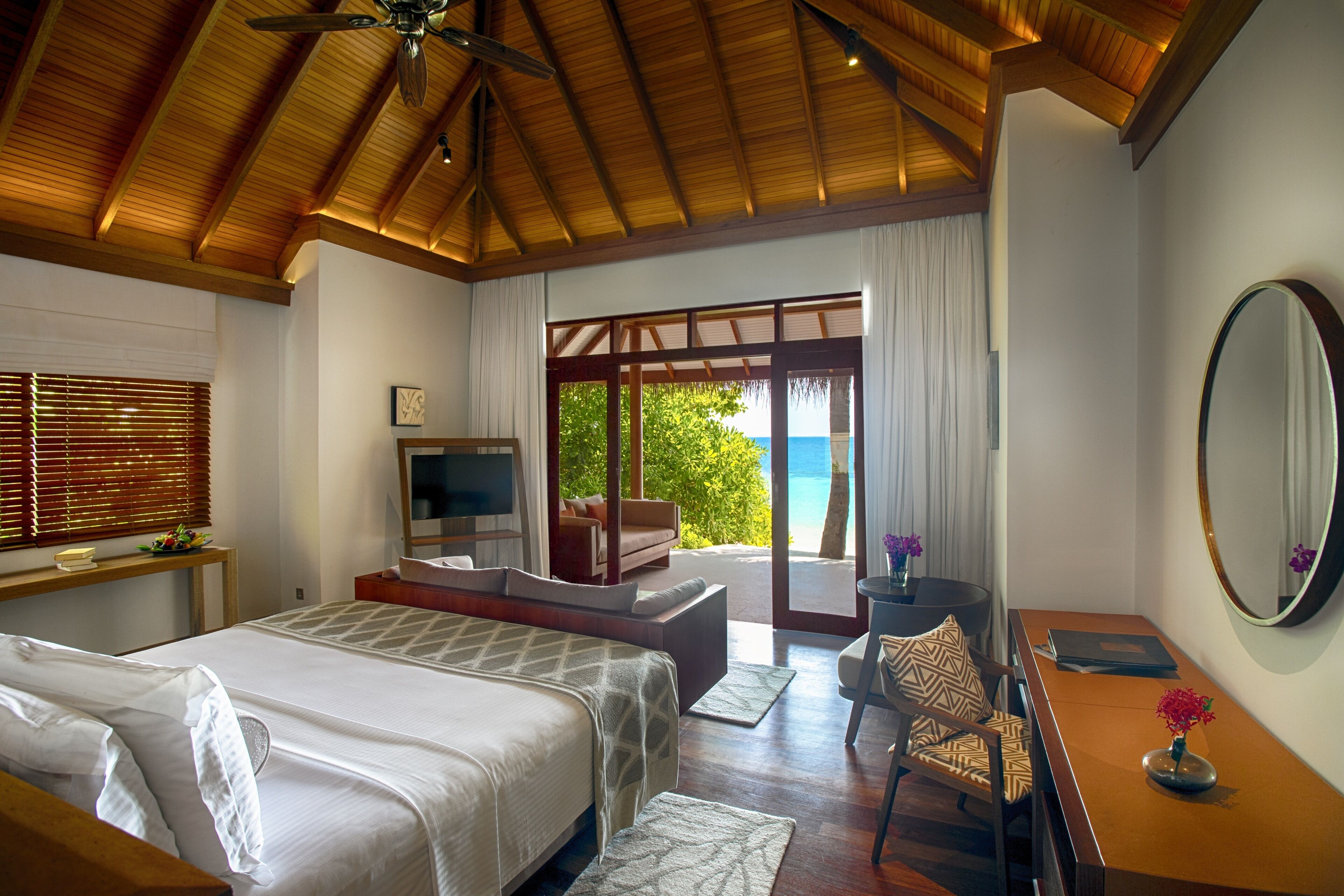 A bedroom with a large bed, a desk, a chair, a TV, and a view of the ocean.