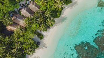 An aerial view of a beach resort with thatched-roof huts, palm trees, and clear turquoise waters.