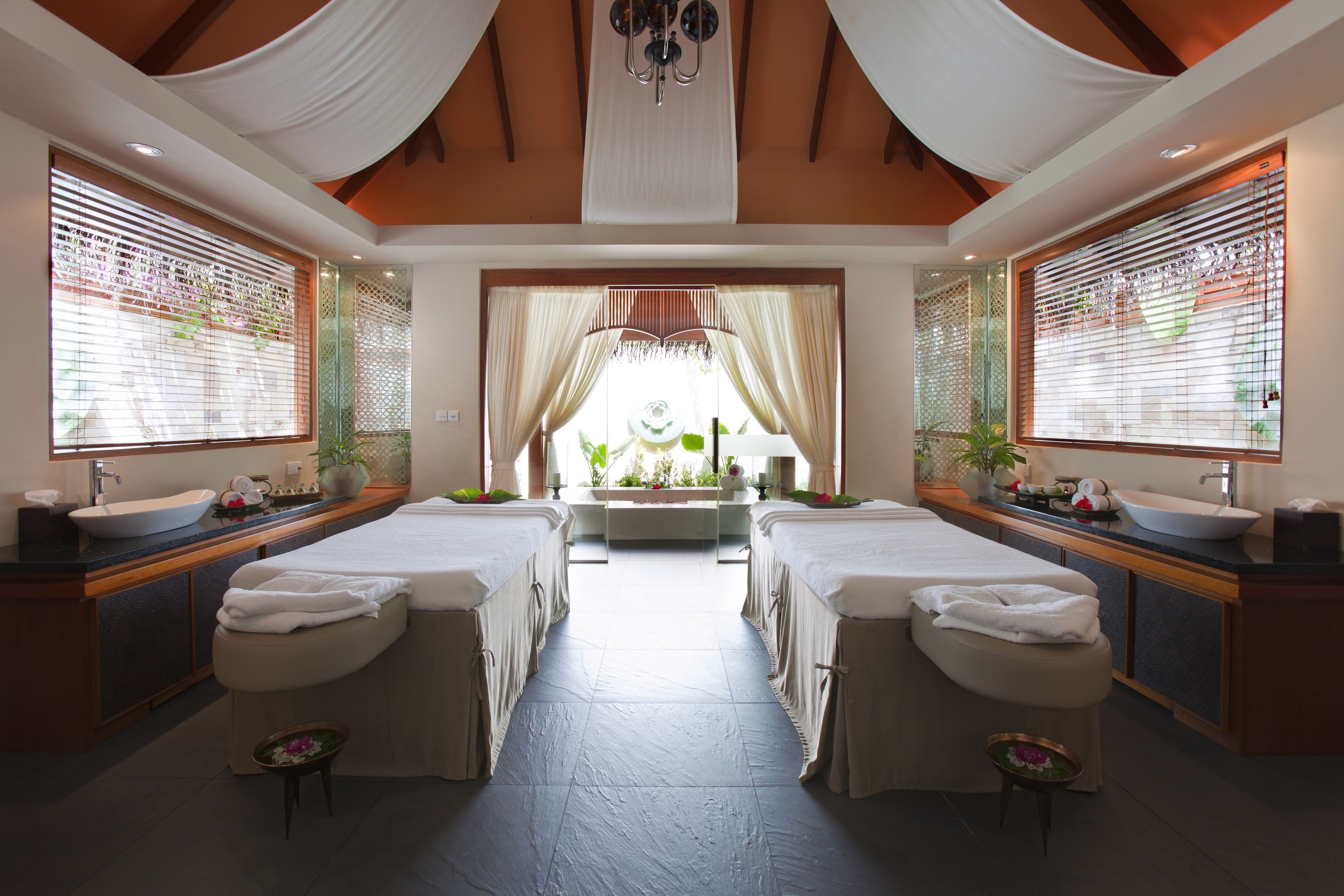 A spa room with two beds, a large window with curtains, a sink, and a mirror.