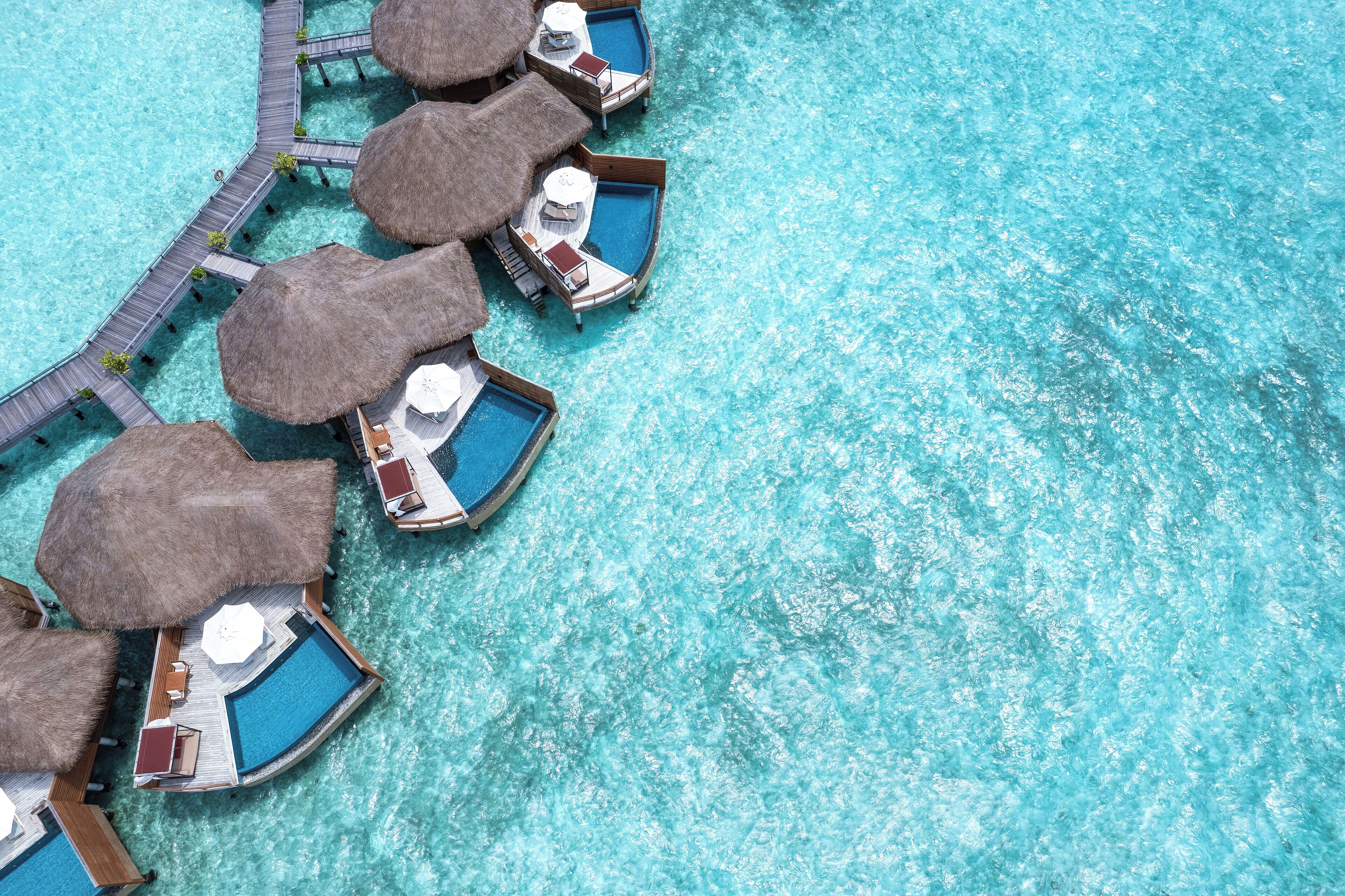 Vue aérienne de bungalows sur pilotis, coiffés de toits de chaume, reliés par une passerelle en bois et entourés d’une eau turquoise et cristalline.