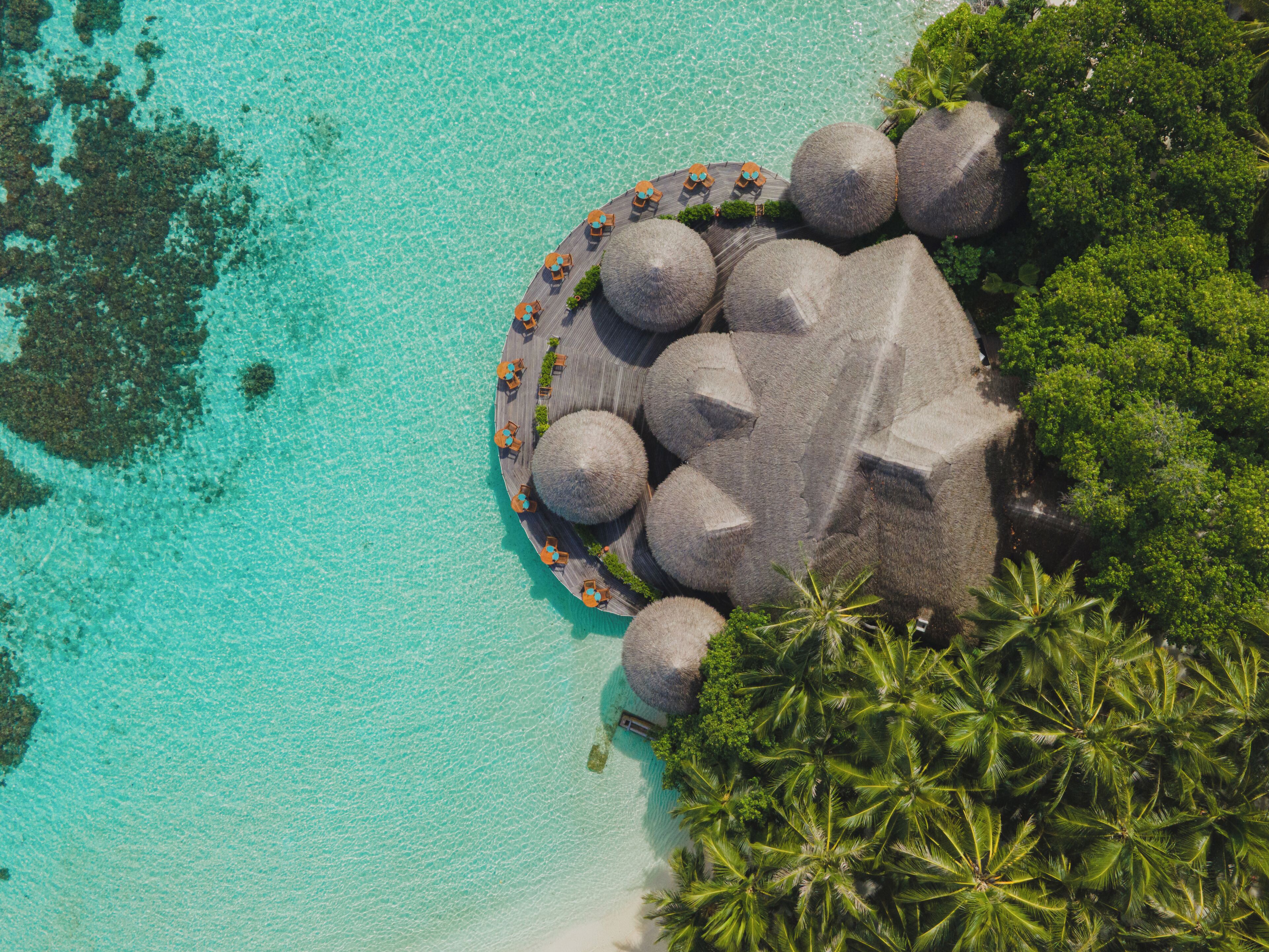 Vista aérea de un complejo turístico con distribución circular, que presenta construcciones con techo de paja y una zona de playa.