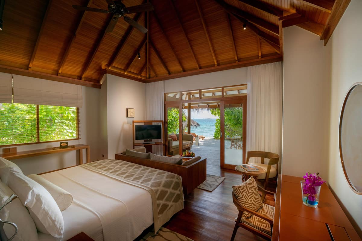 A bedroom with a bed, TV, seating area with a view of the ocean, and a dining table with a vase of flowers.