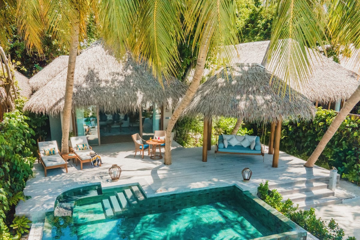 A pool area with lounge chairs and a thatched-roof seating area surrounded by palm trees.