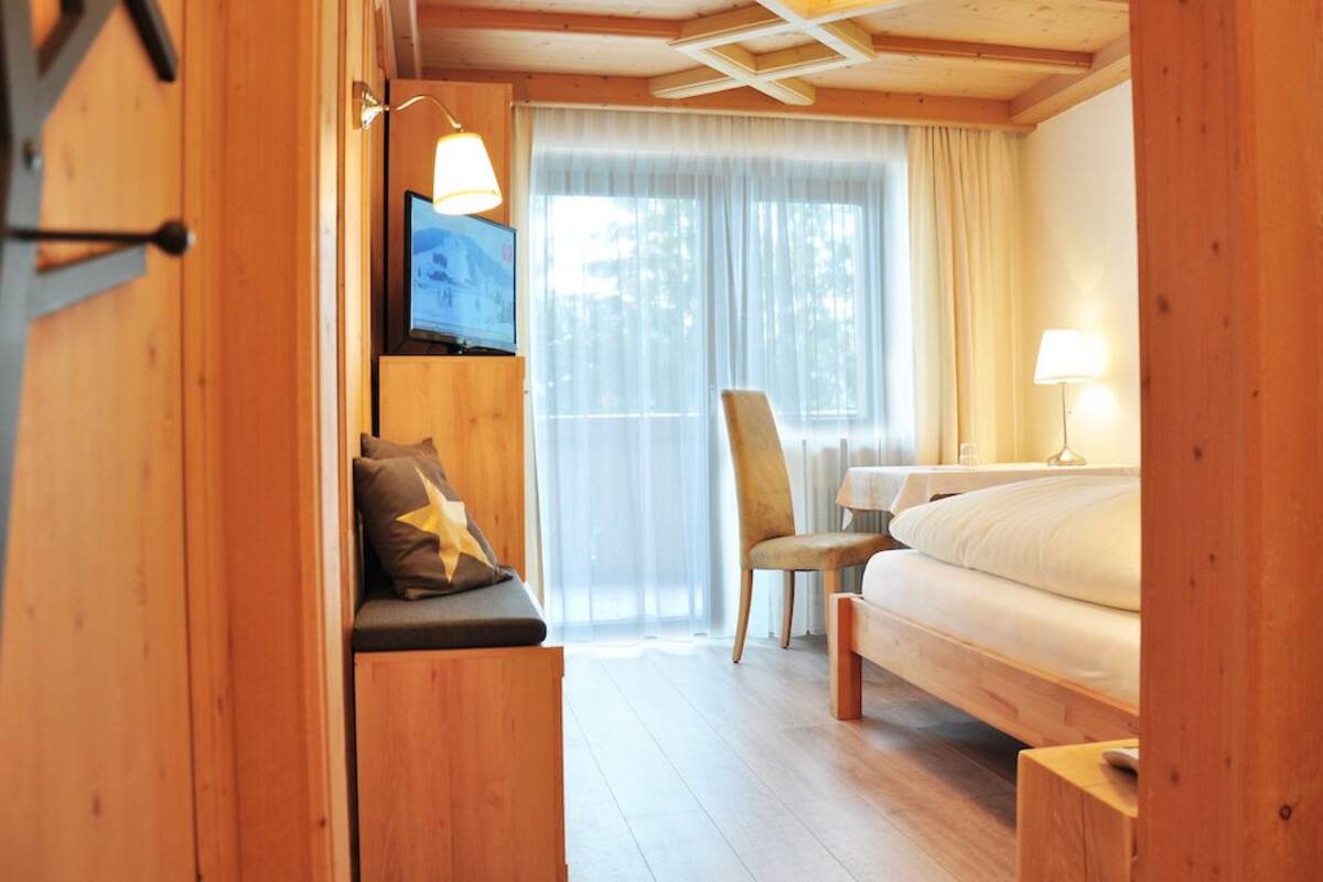 Ein Zimmer in einer Holzhütte mit einem Bett, einem Sessel, einem Schreibtisch mit Fernseher und einem Fenster mit Vorhängen.